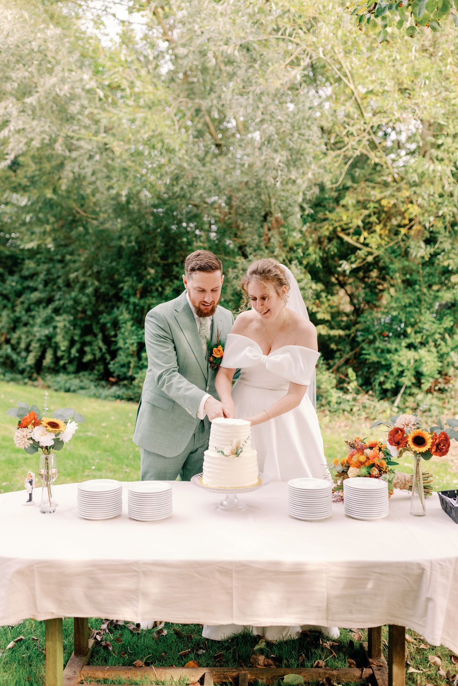 A couple cutting their wedding cake together outdoors at a table decorated with sunflowers and autumn decor.