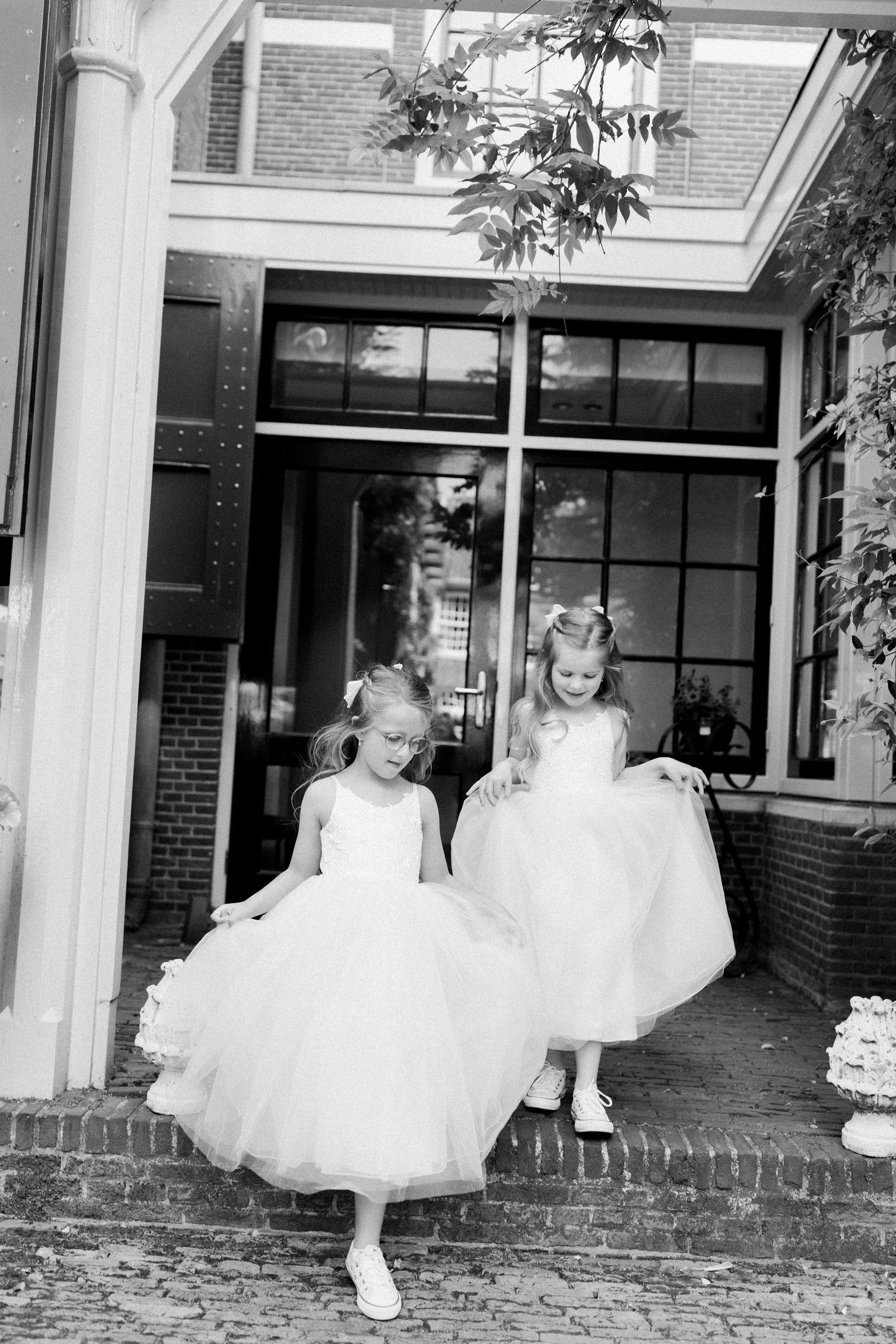 Three young dancers in white tutus practice ballet poses along a covered brick walkway at a dance studio.