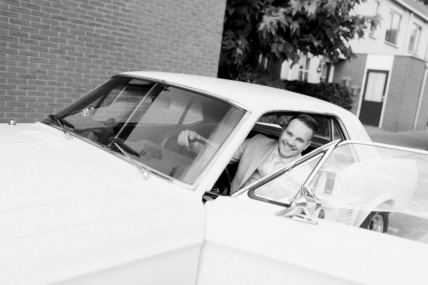 Classic white Chevrolet Corvette convertible parked on street next to brick building with other vintage cars visible.