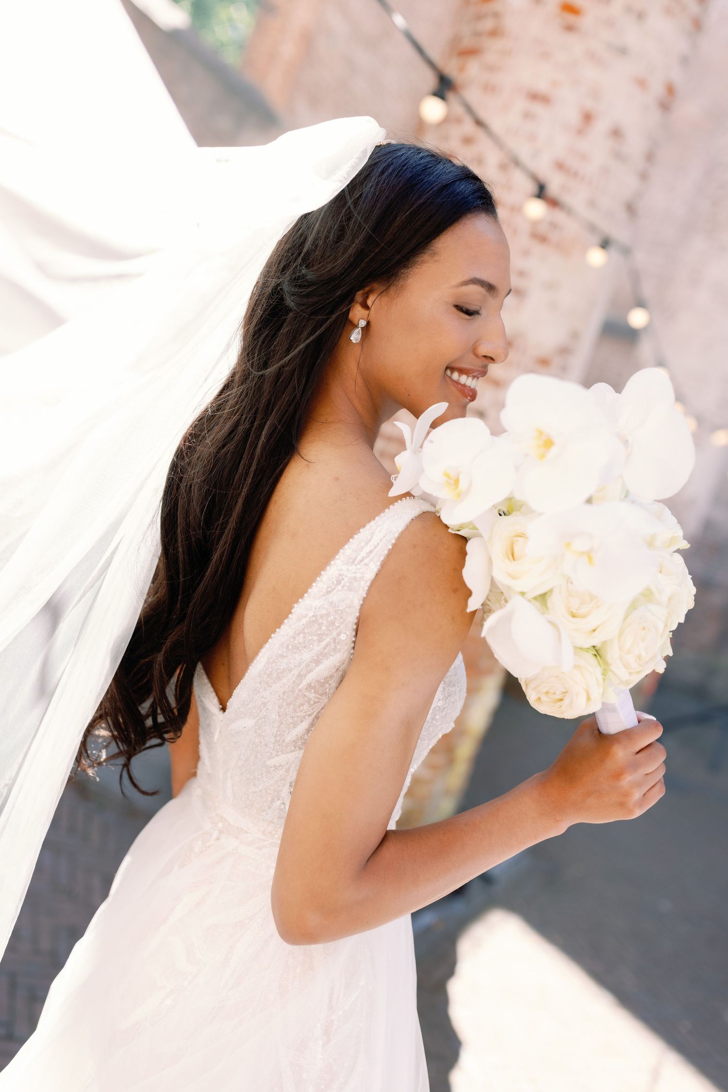 A bride in a white lace dress smiles while holding a bouquet of white flowers in a romantic outdoor setting.