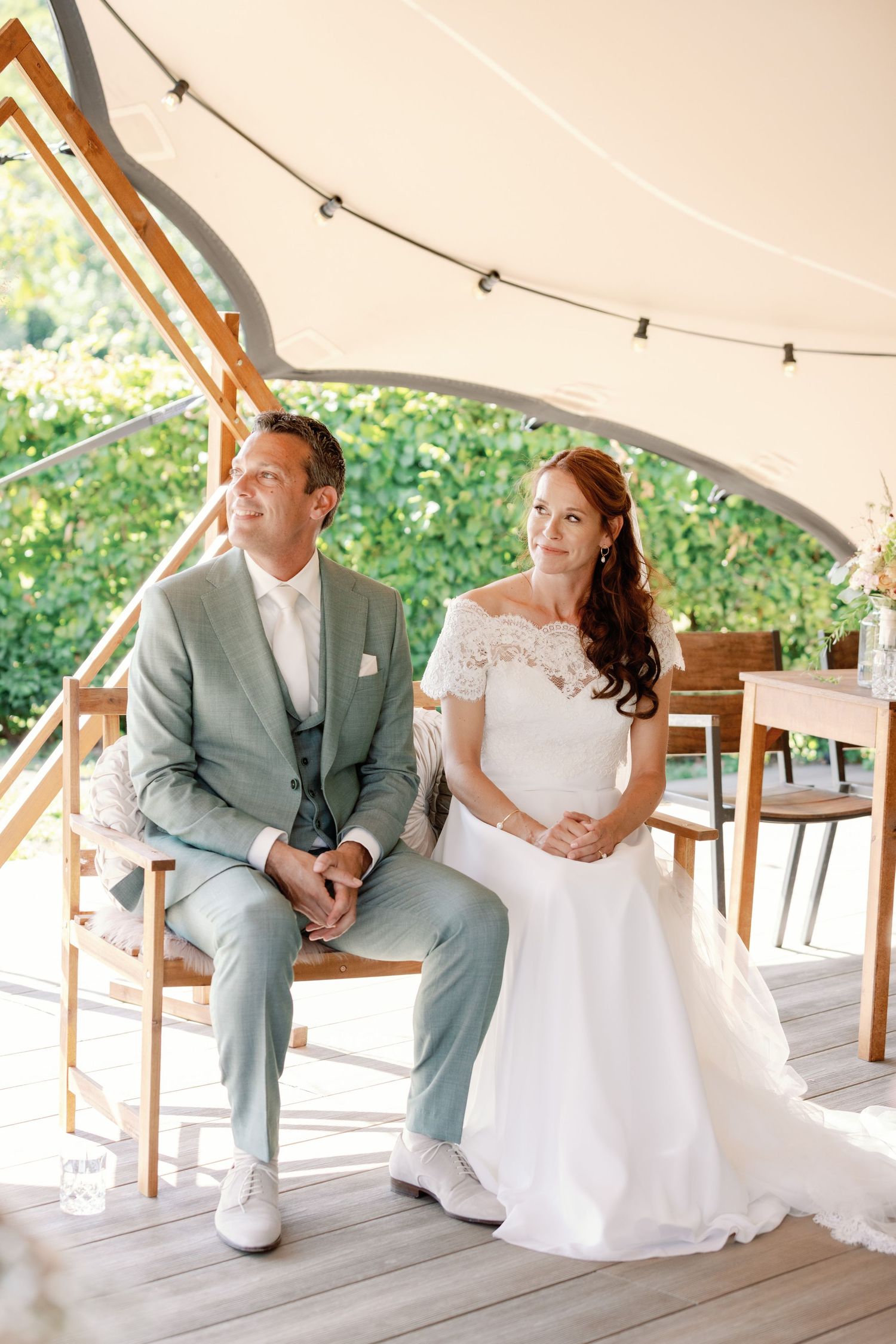 A couple in wedding attire sits on a white bench under a tent canopy during their outdoor celebration.