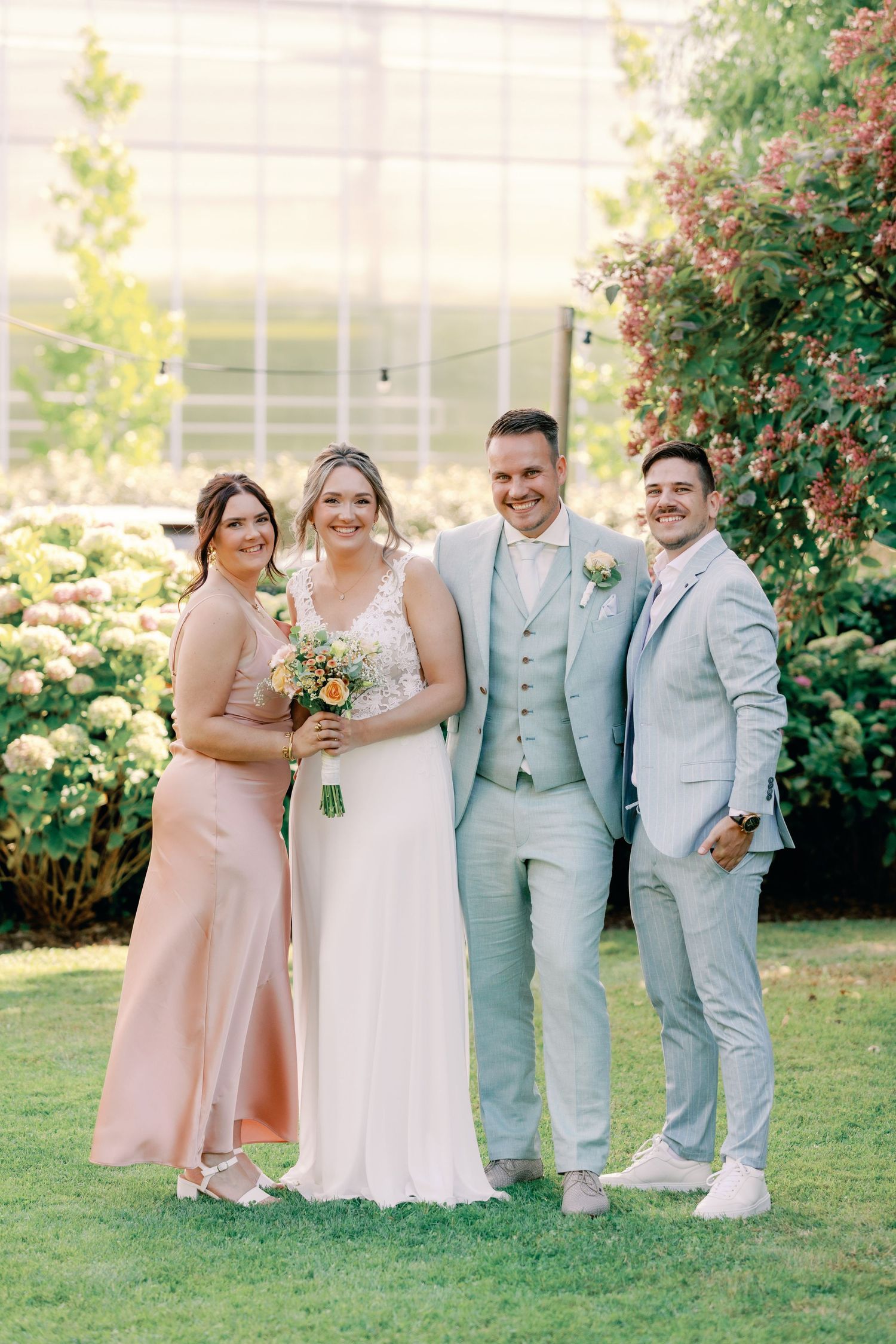 Wedding party in pastel pink and mint green attire poses together on a sunny lawn with blooming flowers.