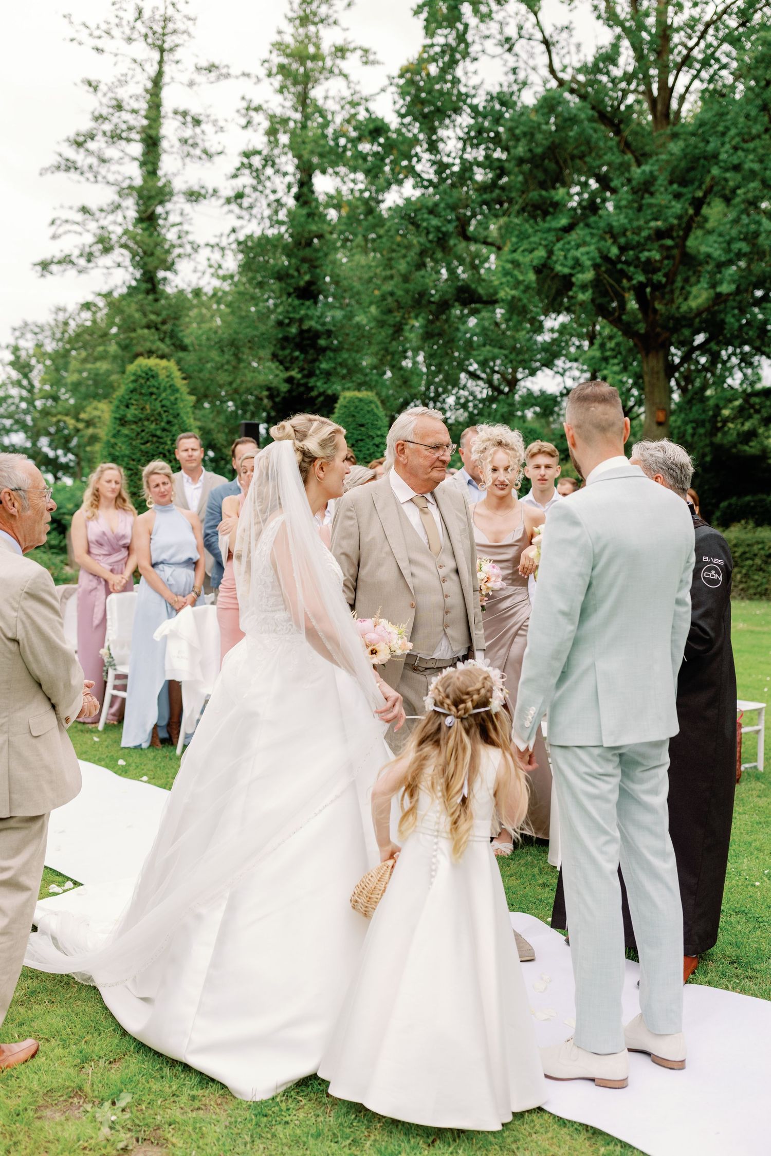 Outdoor garden wedding ceremony with guests gathered around a white aisle runner surrounded by lush green trees.