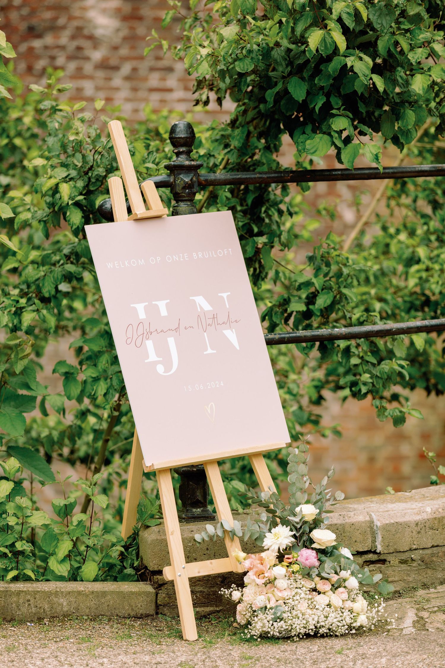 A pink wedding sign on a wooden easel stand with delicate floral decorations against a brick wall and greenery background.