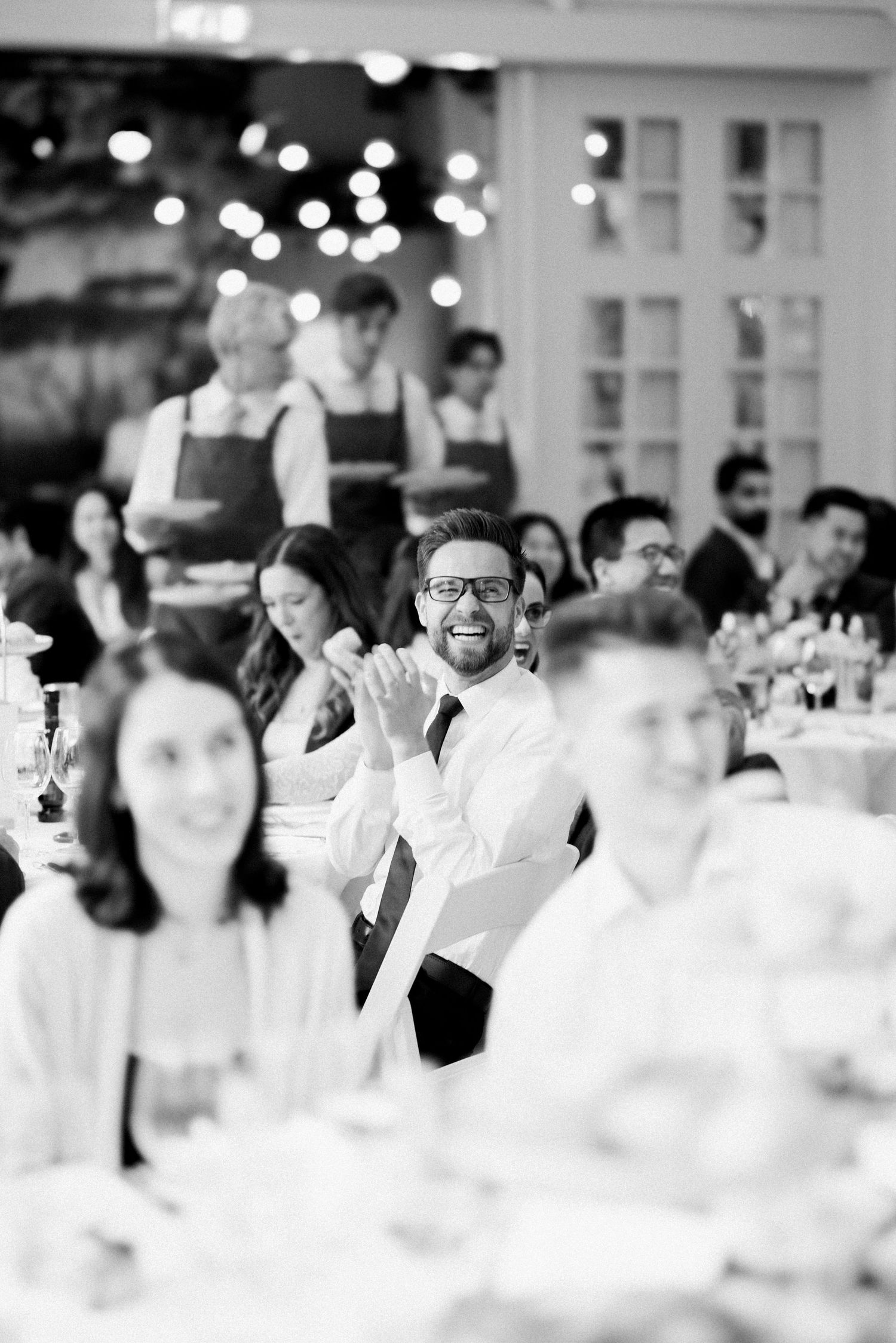 Black and white photo of wedding reception guests enjoying a celebration with bokeh lights and white decor in background.
