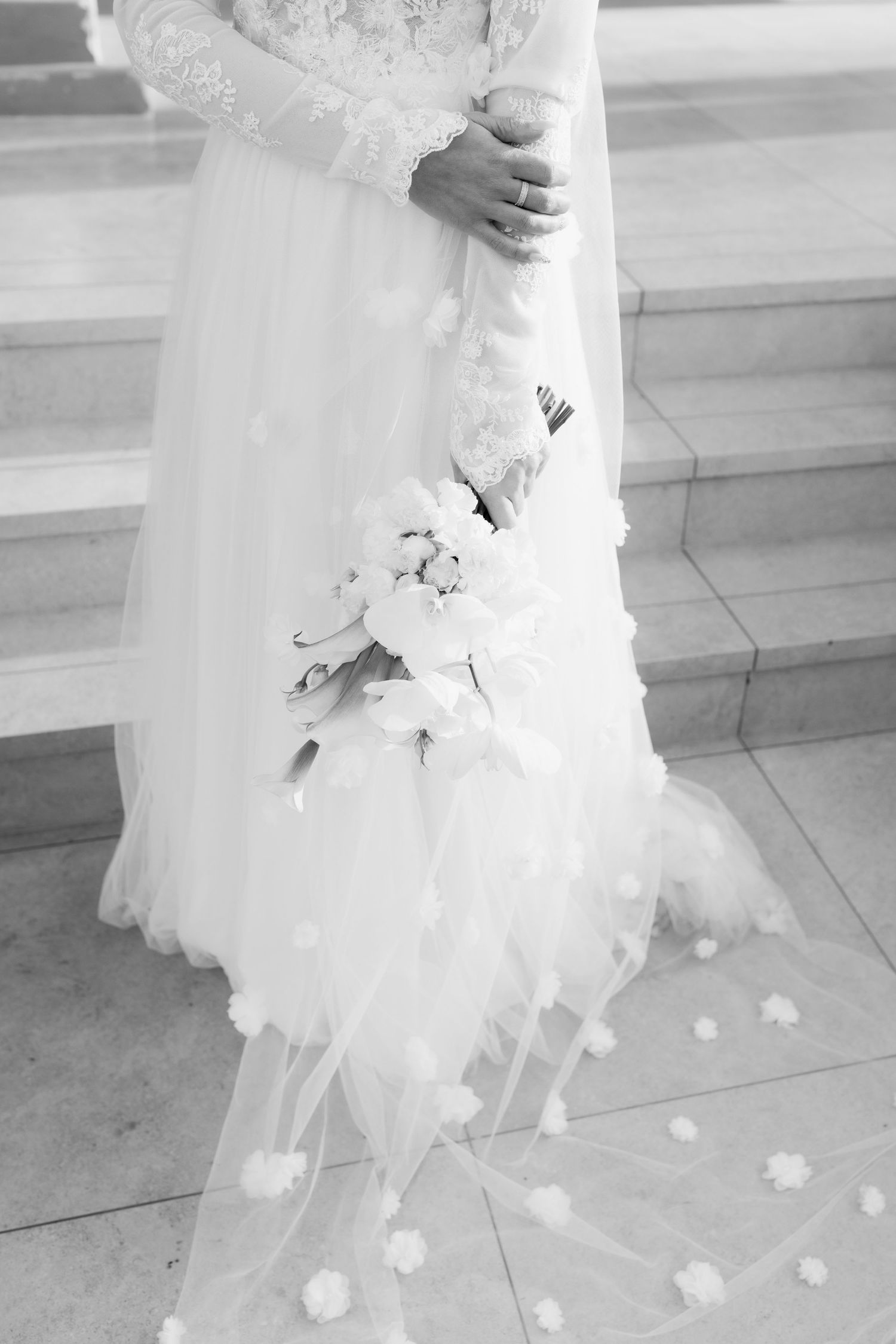 The bottom of a white wedding dress with delicate floral details and scattered rose petals on a marble floor.