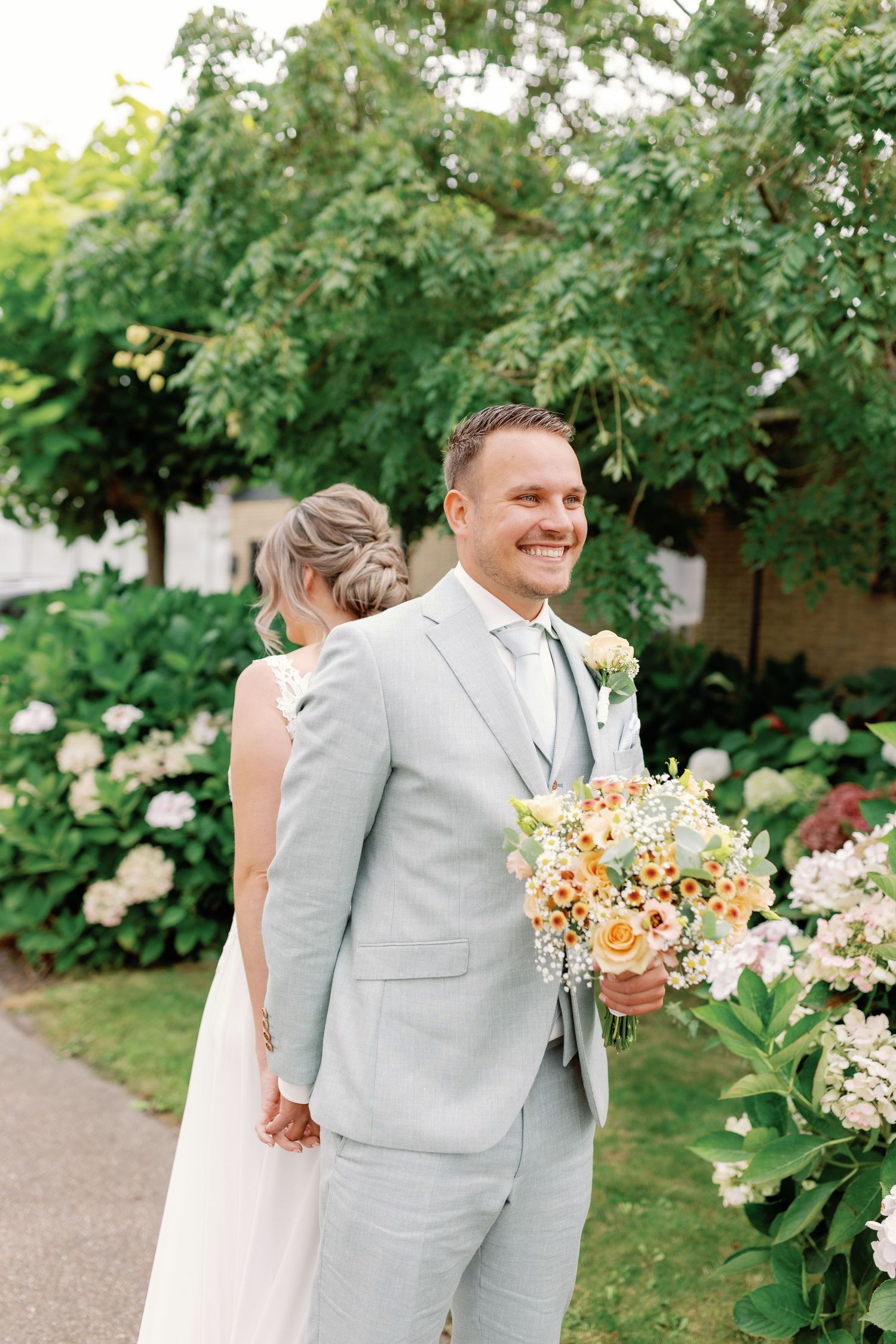 A couple in wedding attire walking together through a lush garden path with peach-colored floral bouquet.