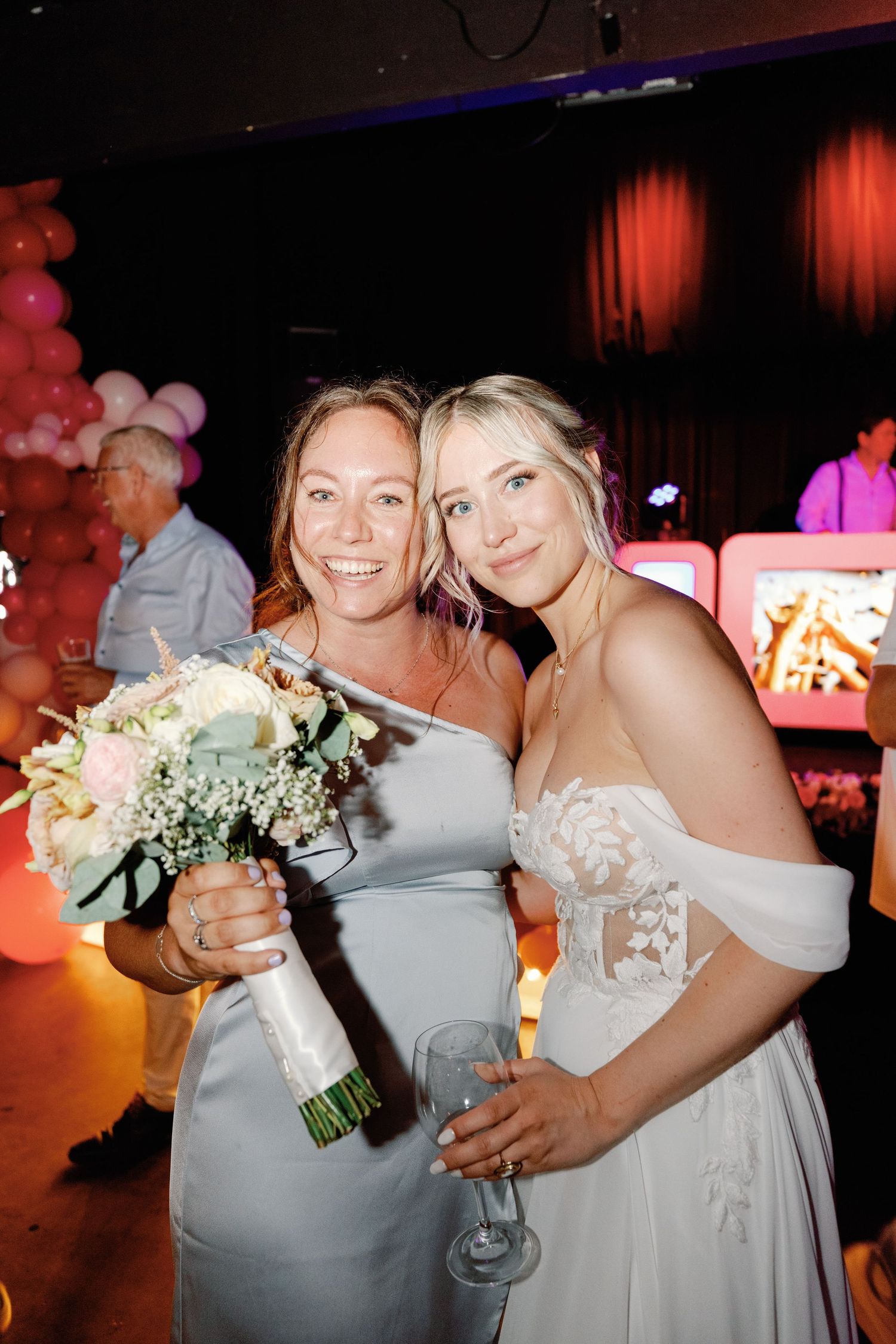 Wedding party guests in light-colored formal attire dance and celebrate at a reception with red ambient lighting.