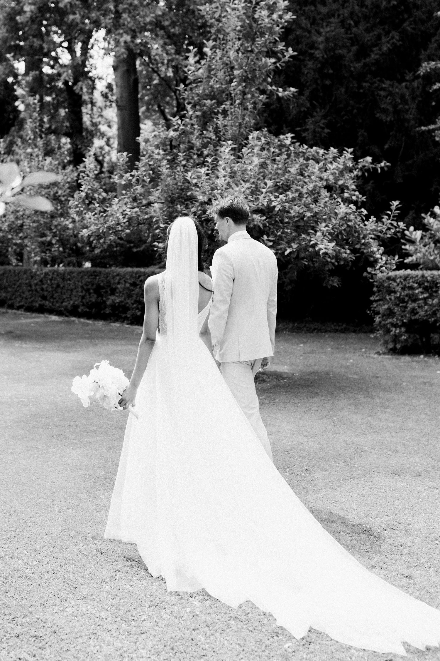 A black and white wedding photograph shows a couple walking together on a garden lawn with a flowing white dress trailing behind.