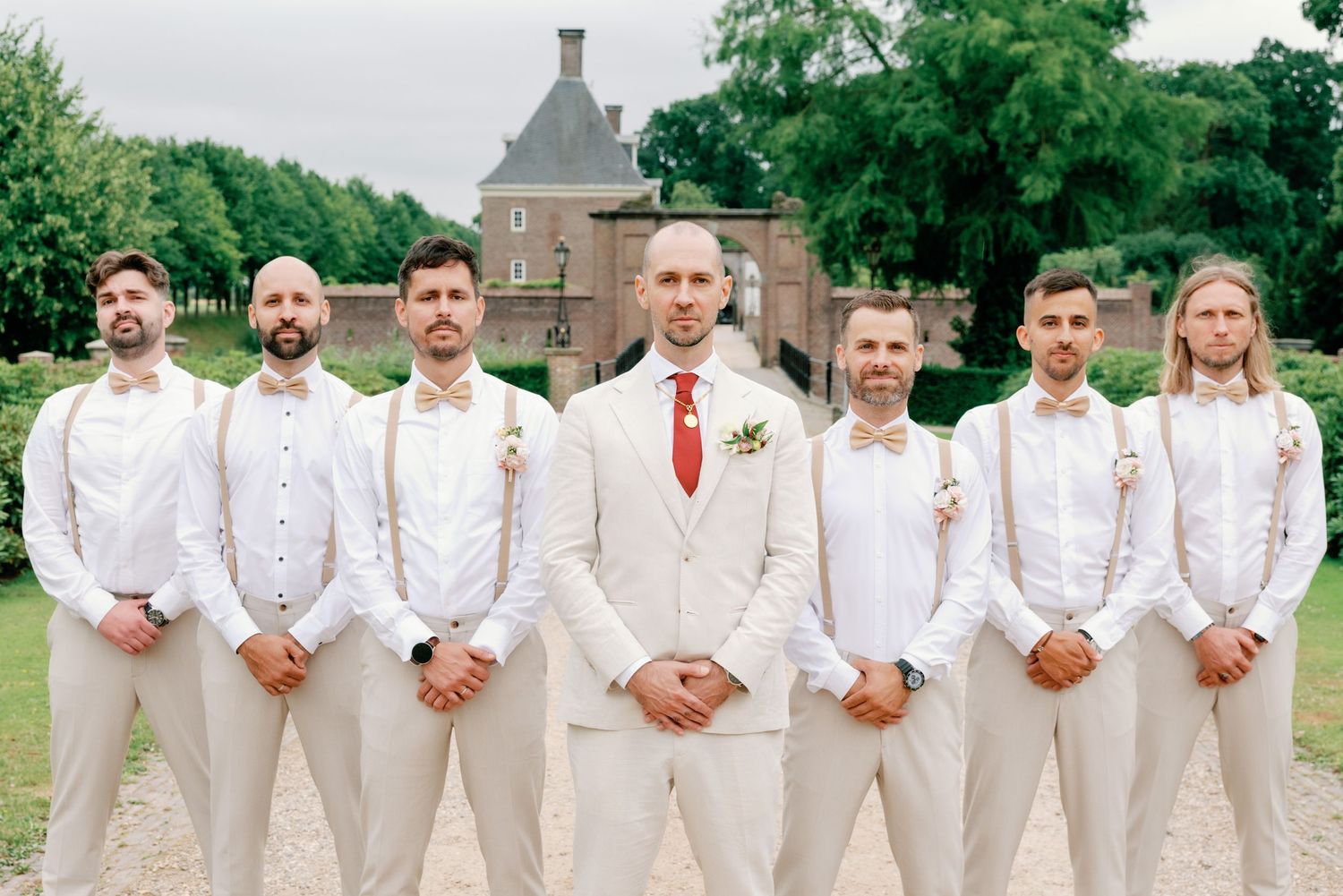 A wedding party wearing white suits and red ties poses together outdoors on a grassy lawn.