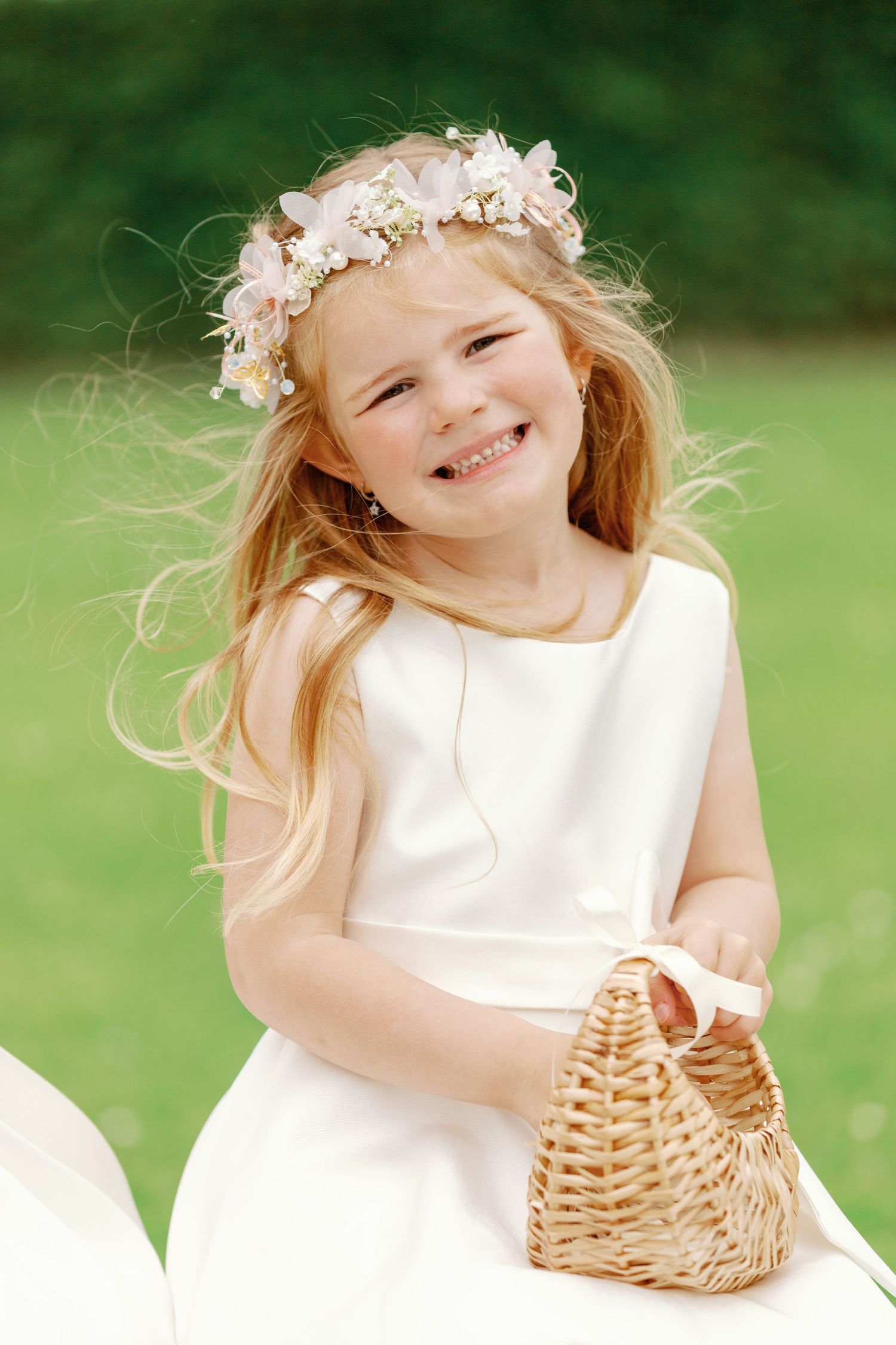 A flower girl in a white dress holds a wicker basket while standing outdoors in a garden setting.