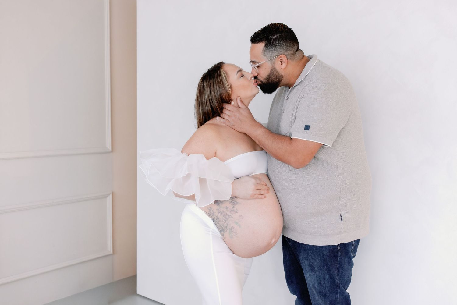 A series of intimate maternity photos showing a couple in casual attire against a white wall.