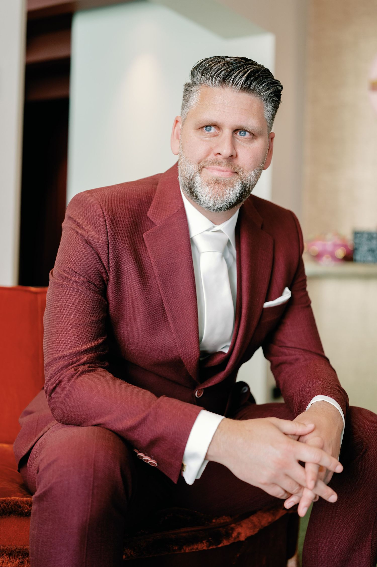 Professional portrait of someone in a burgundy suit and white tie sitting on a red couch in an elegant setting.