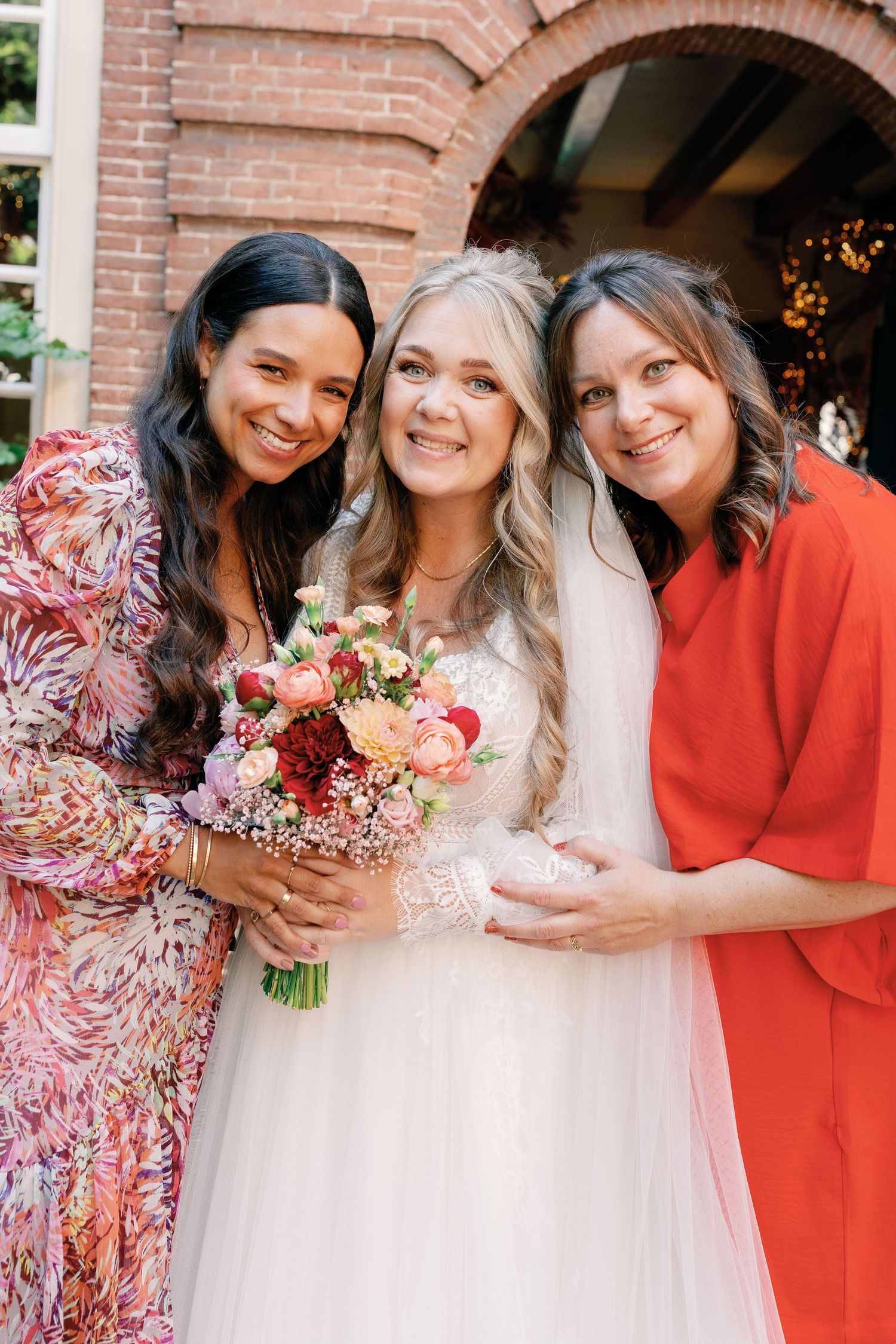 A bridal party poses together wearing white and coral dresses with pink floral bouquets against a brick wall.