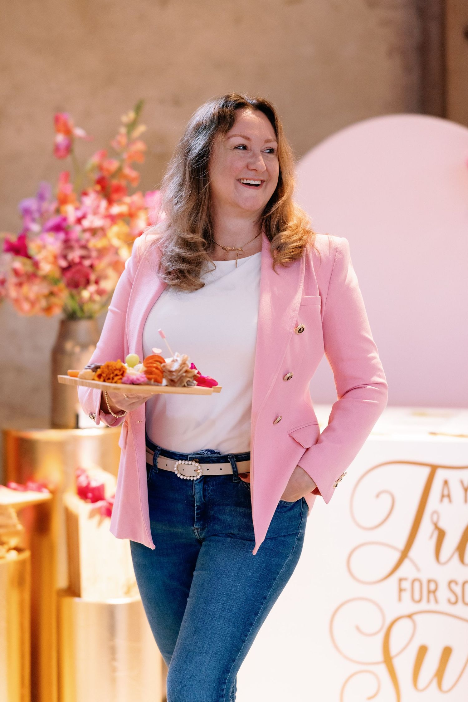 A woman in a pink blazer and jeans smiles while standing in front of a floral display and gold decor.