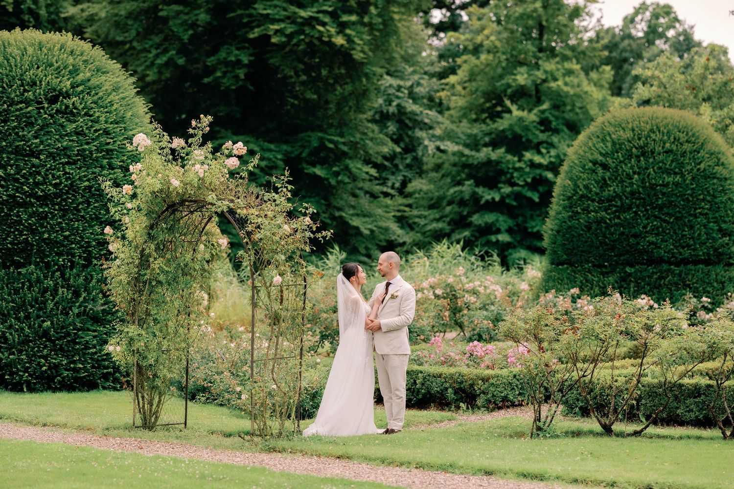 A couple in wedding attire embraces in a lush garden with manicured hedges and blooming flowers on a summer day.