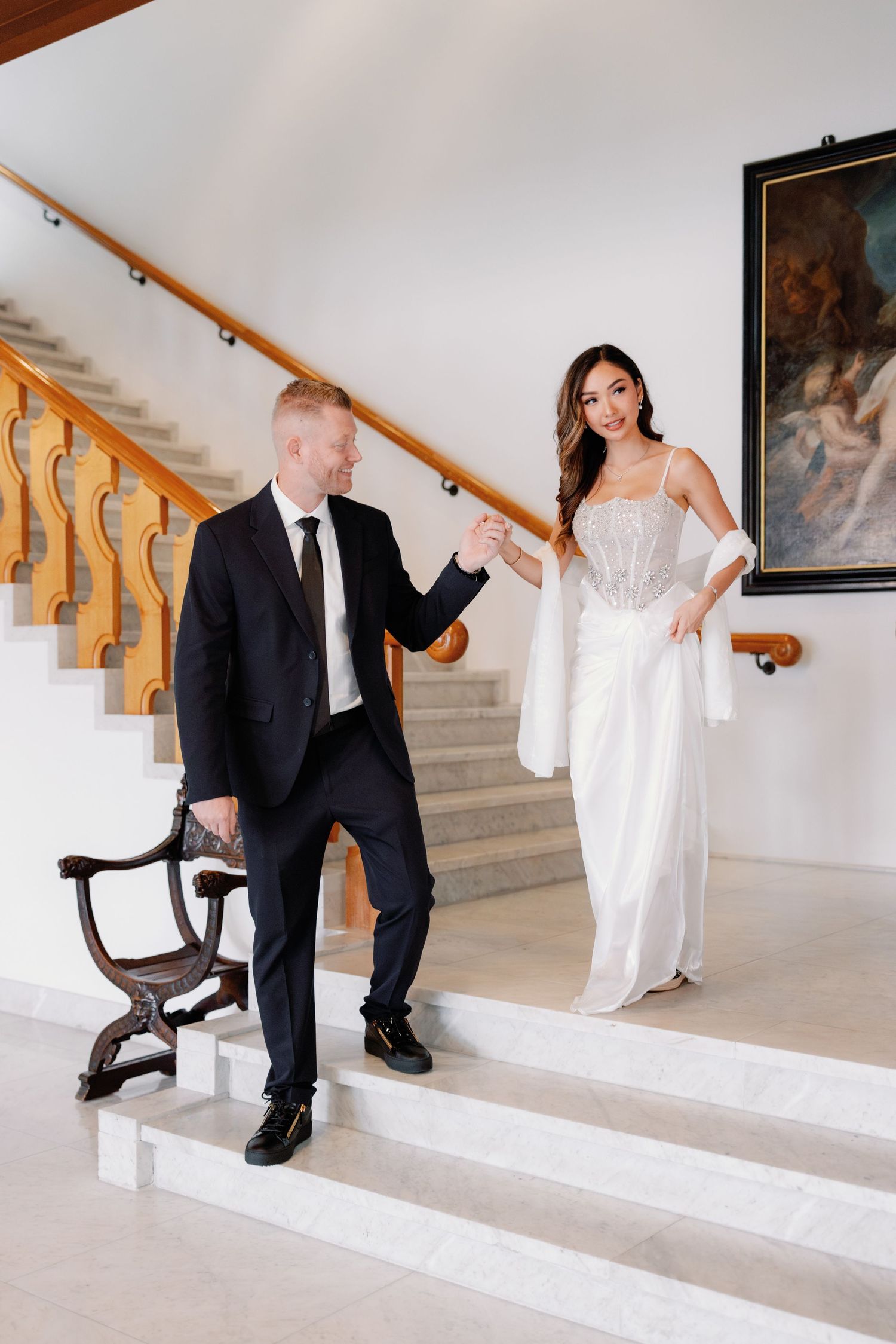Bride in a white gown holds the hand of a groom in a dark suit as they stand on marble steps beside a wooden staircase.