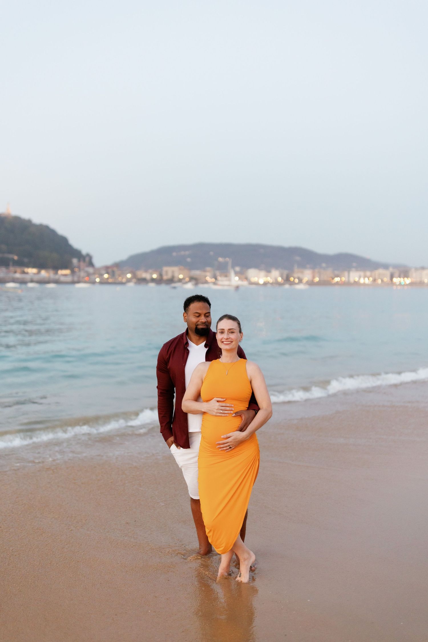 Couple sharing a romantic moment on a sandy beach at sunset with ocean and mountains in background.