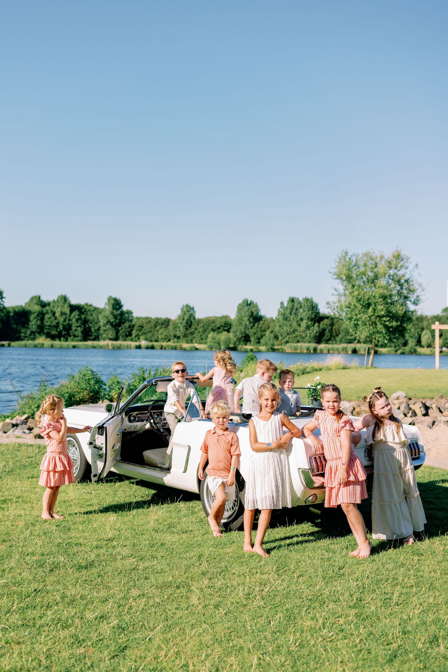 Wedding party lounges on vintage couches arranged on a grassy lawn near a lake on a sunny day.
