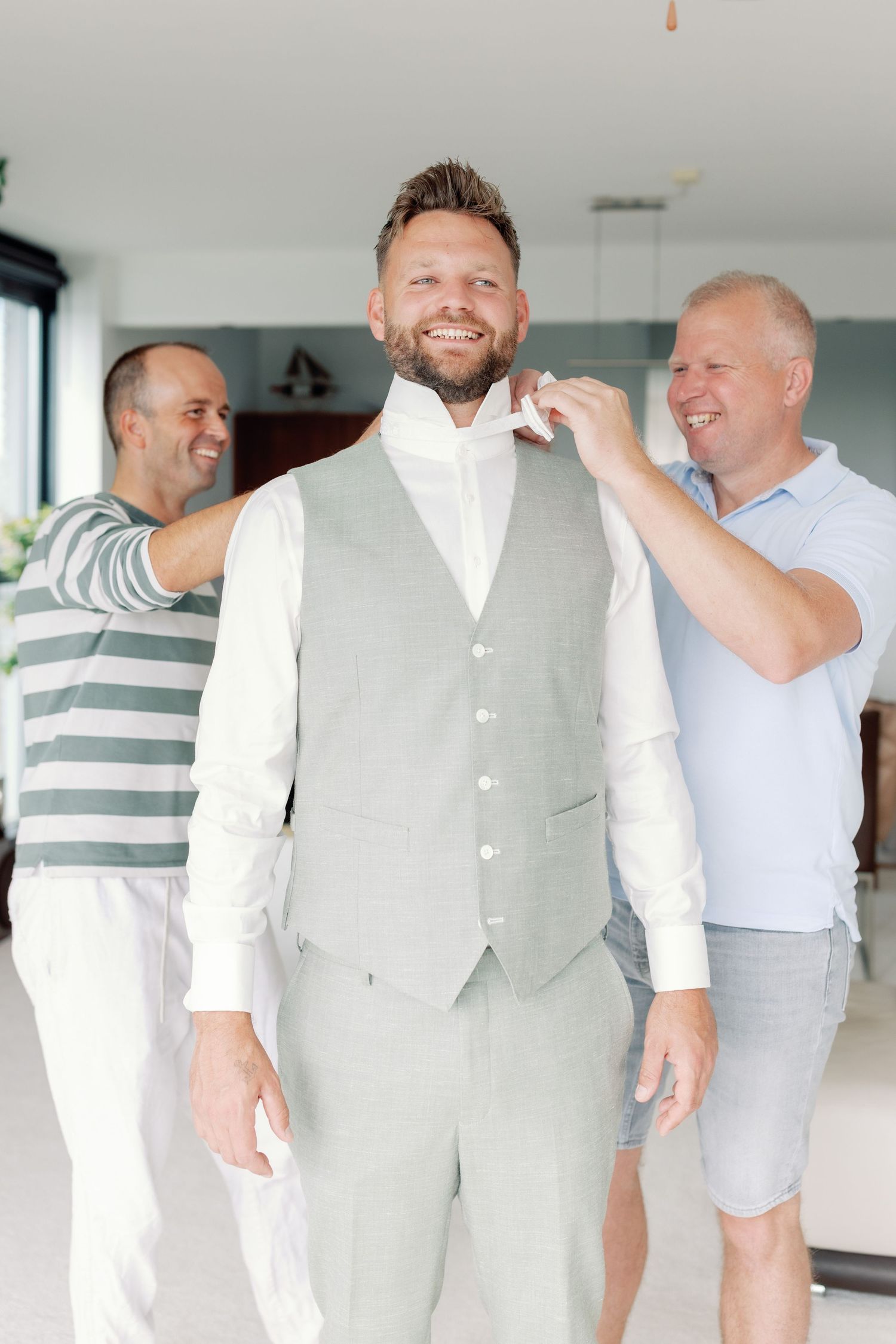 Two people help adjust a light gray vest on someone getting dressed in formal attire for a special occasion.