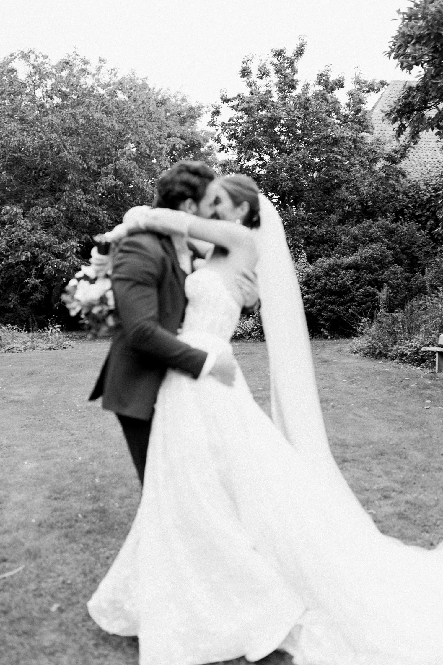 A couple in wedding attire dances and spins together in an outdoor garden setting with trees in the background.