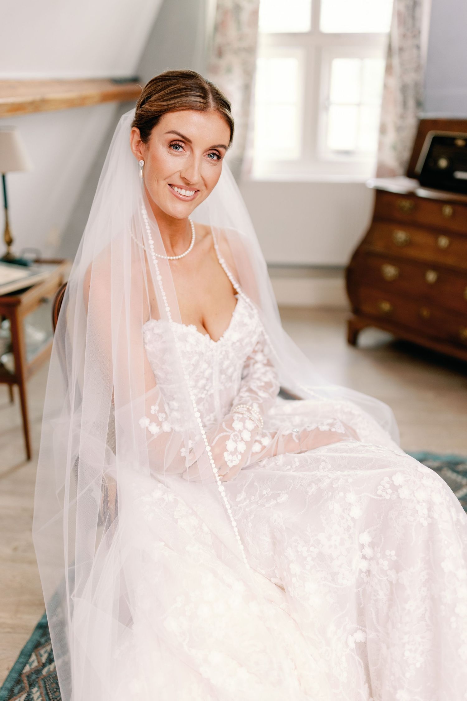 A bride wearing a romantic lace wedding gown sits elegantly in a bright, sunlit room with white decor.