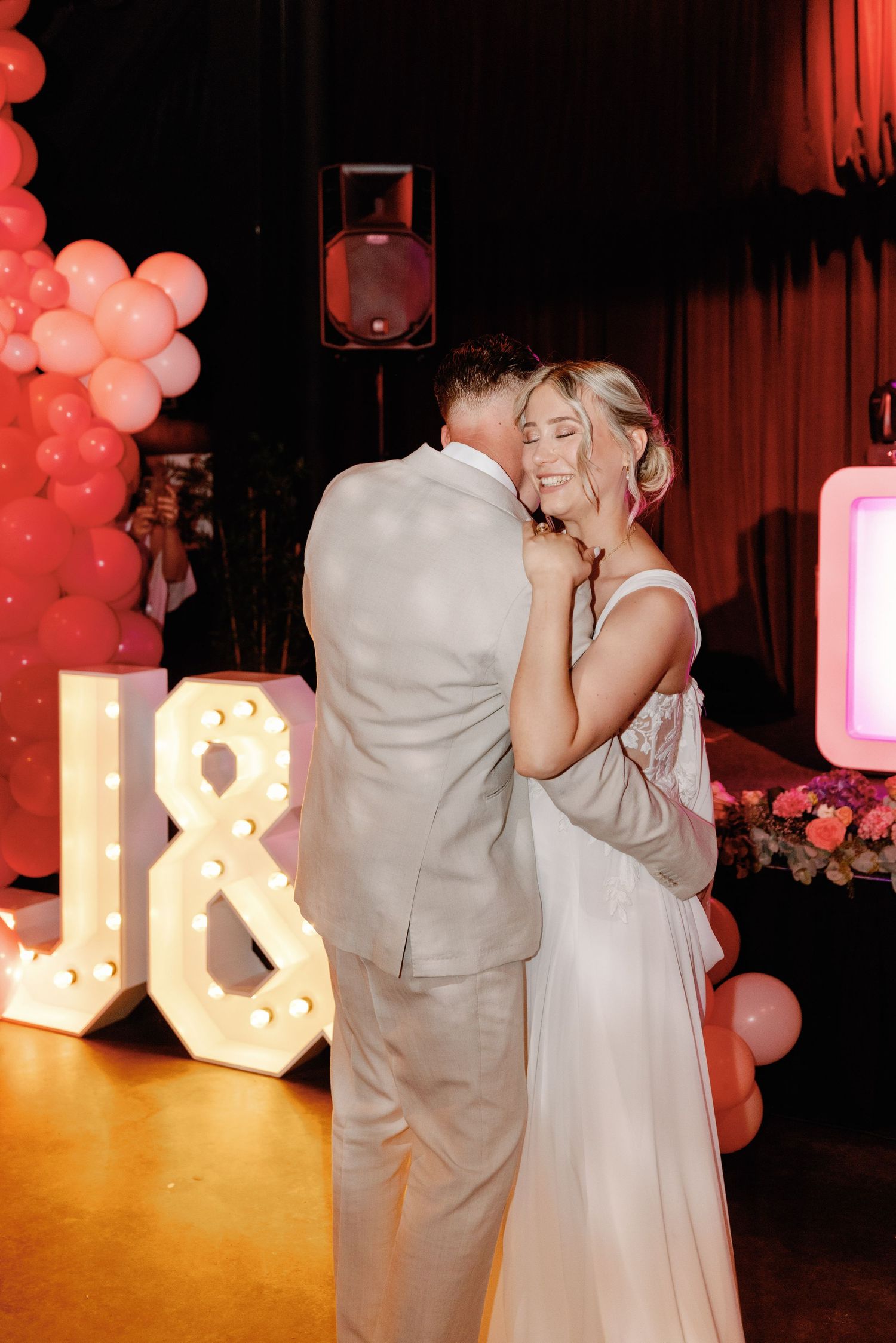 A couple shares a romantic dance at a wedding reception with illuminated letters and pink balloons in the background.