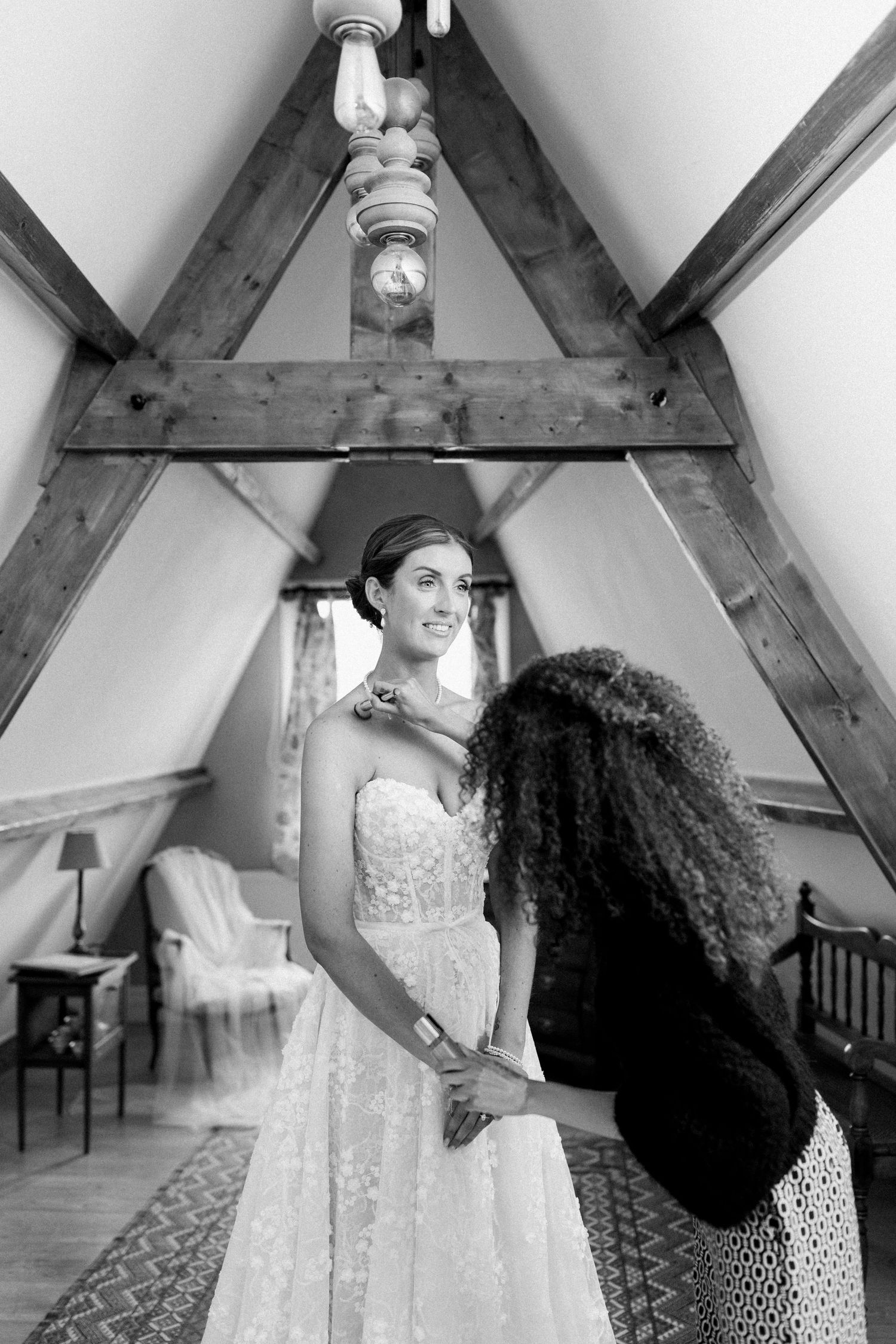 A wedding dress fitting session showing adjustments being made to a white lace gown in an attic room with exposed beams.