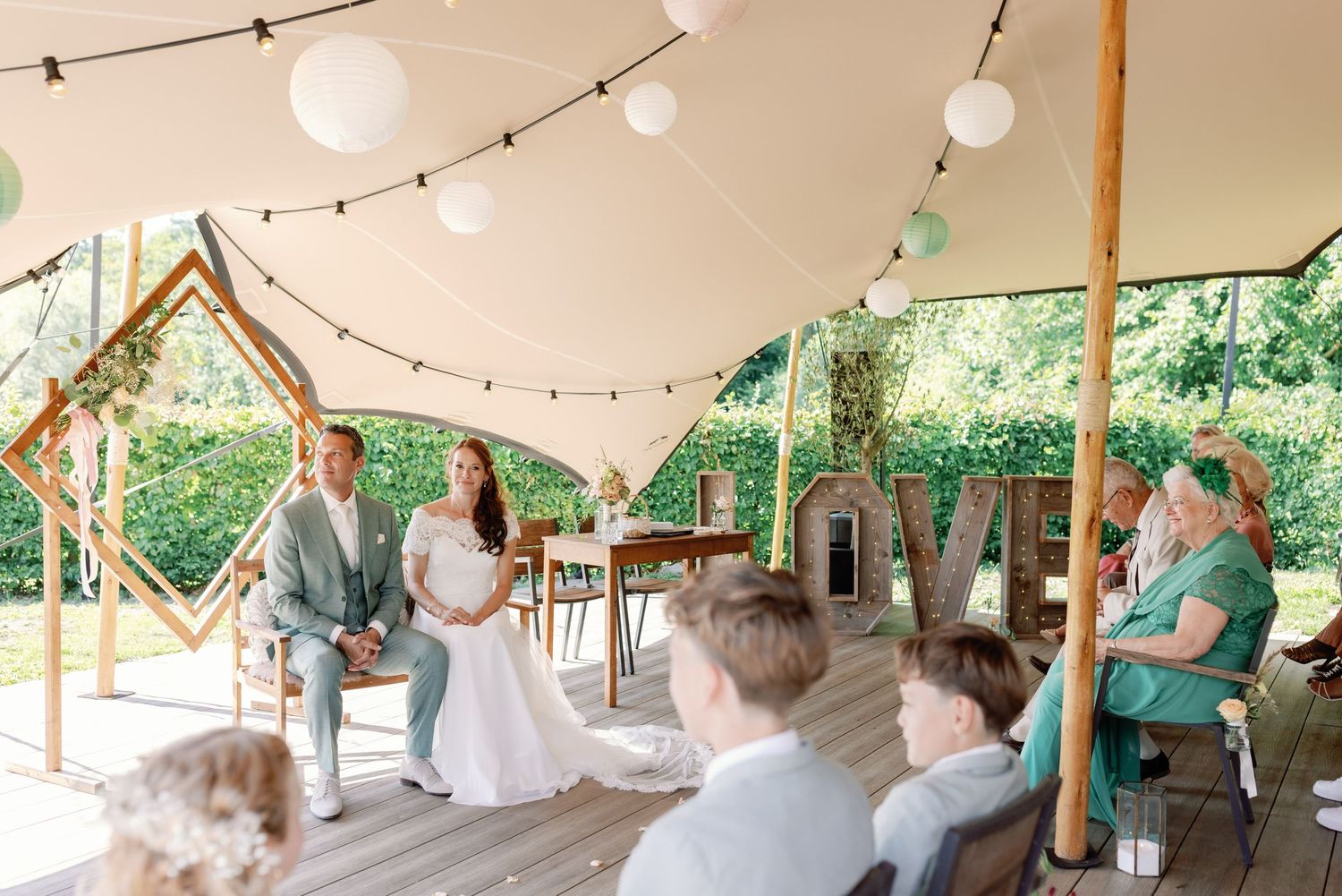 Guests seated at tables under outdoor tent with string lights at rustic wedding reception.