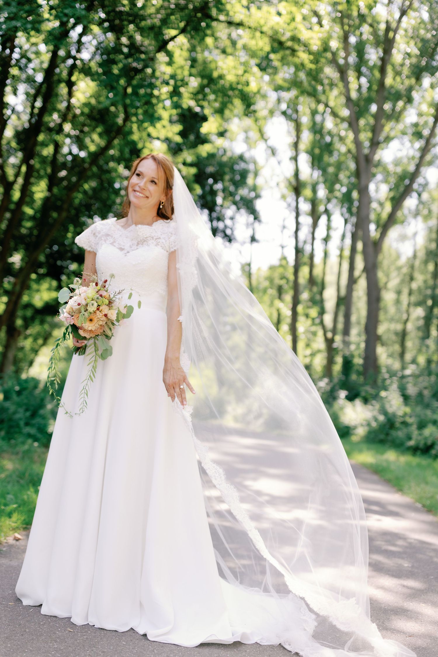 A bride in a flowing white wedding gown twirls gracefully on a sunlit forest path, her veil floating in the breeze.