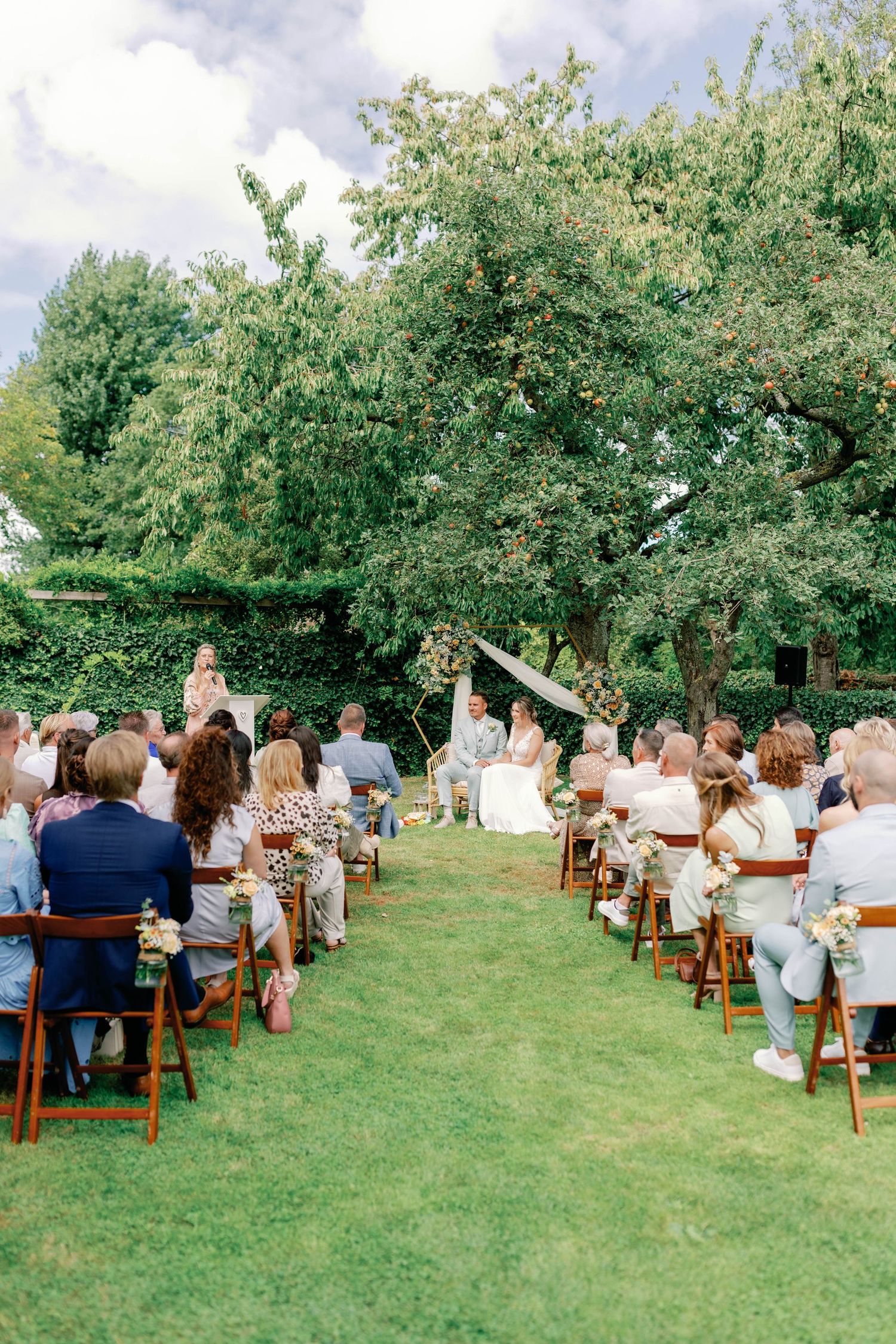 Guests seated in wooden chairs on a lush green lawn during an outdoor garden ceremony or event.