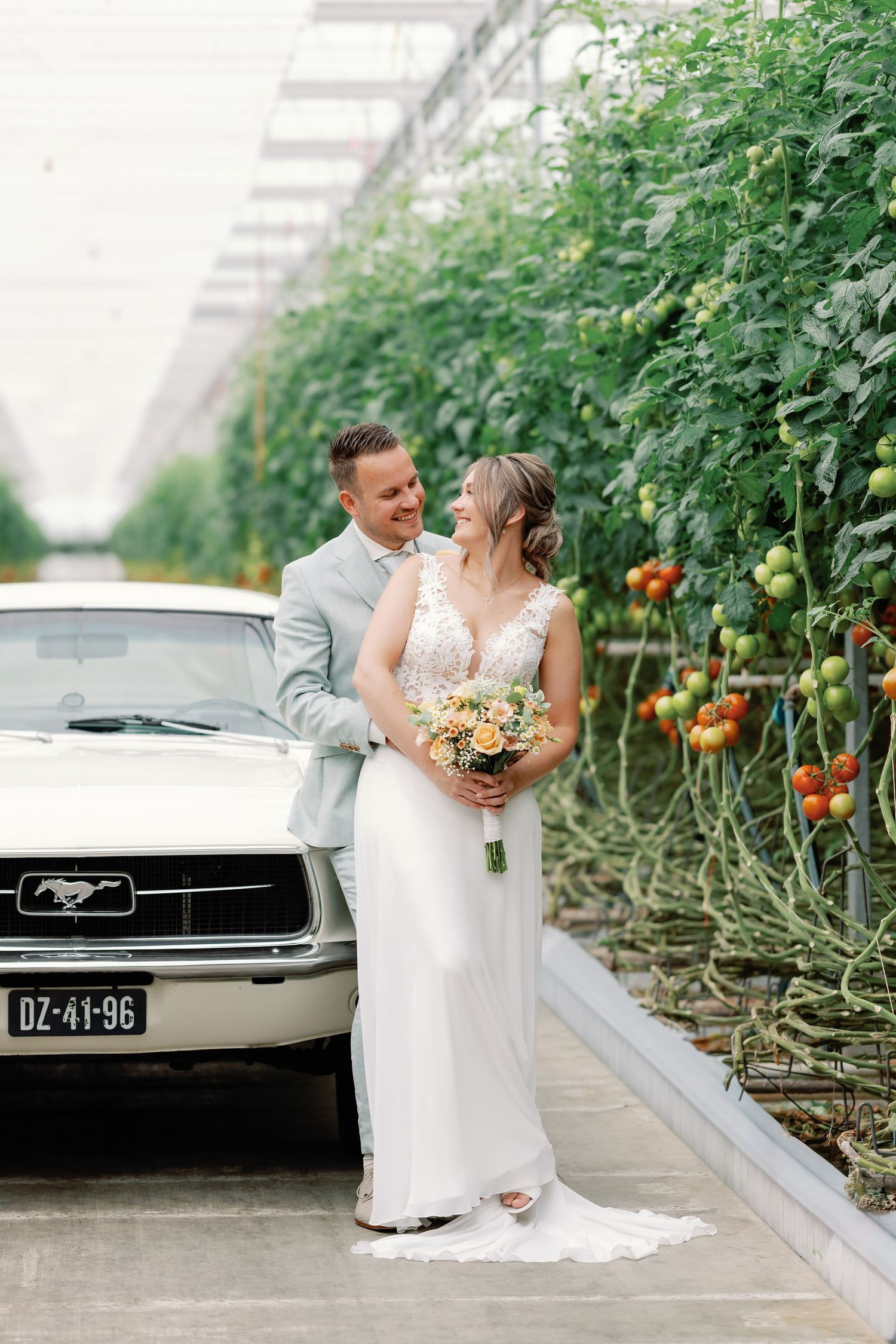 Newlywed couple sharing romantic moments in a greenhouse with tomato plants and a classic Ford Mustang parked outside.
