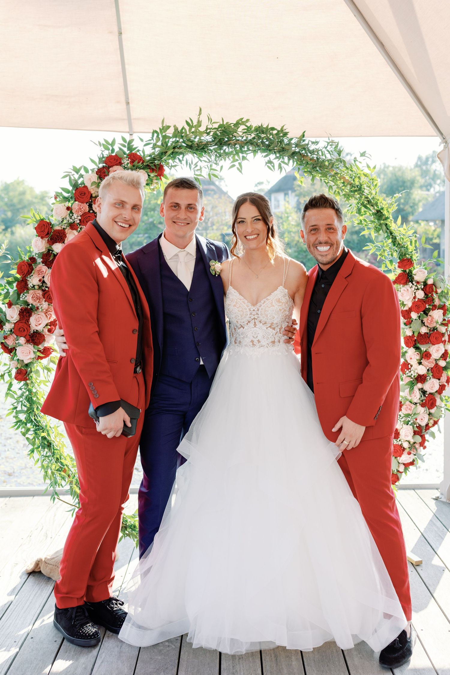 Wedding party poses under floral arch with bride in white gown and groomsmen in red suits.