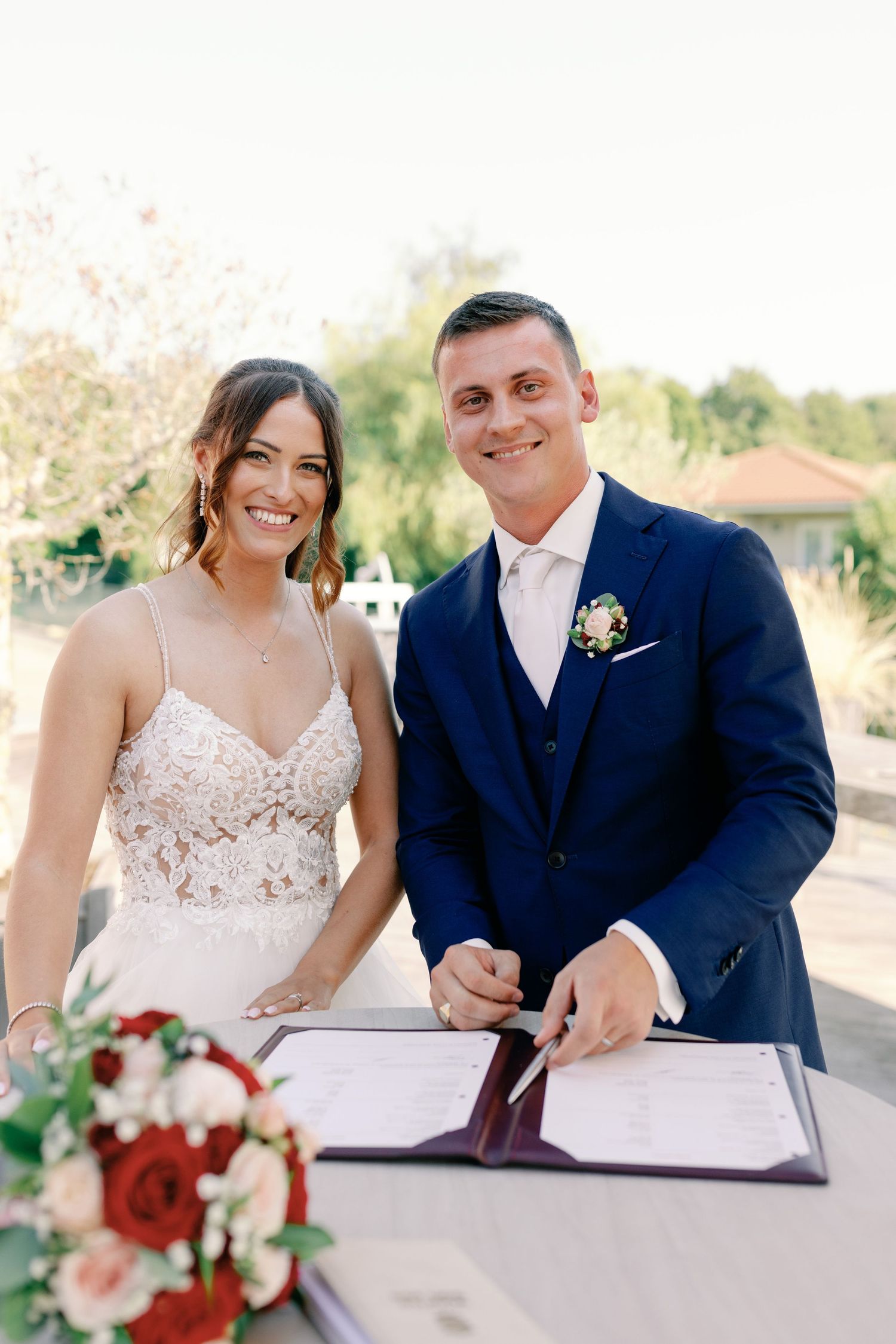 Couple signs marriage documents at outdoor ceremony with floral decorations.