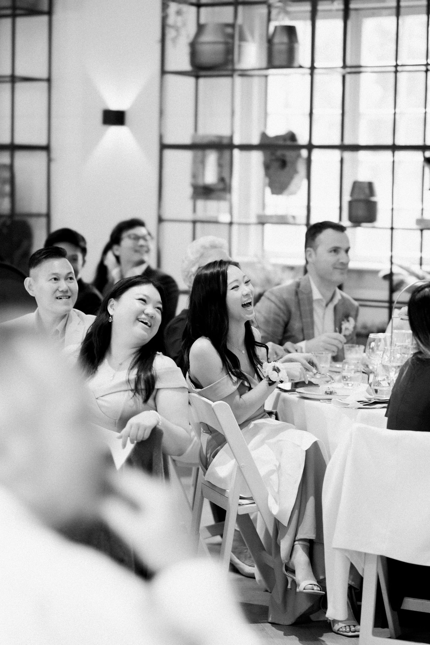 Wedding guests laugh and share joyful moments during reception dinner at tables in bright windowed venue.