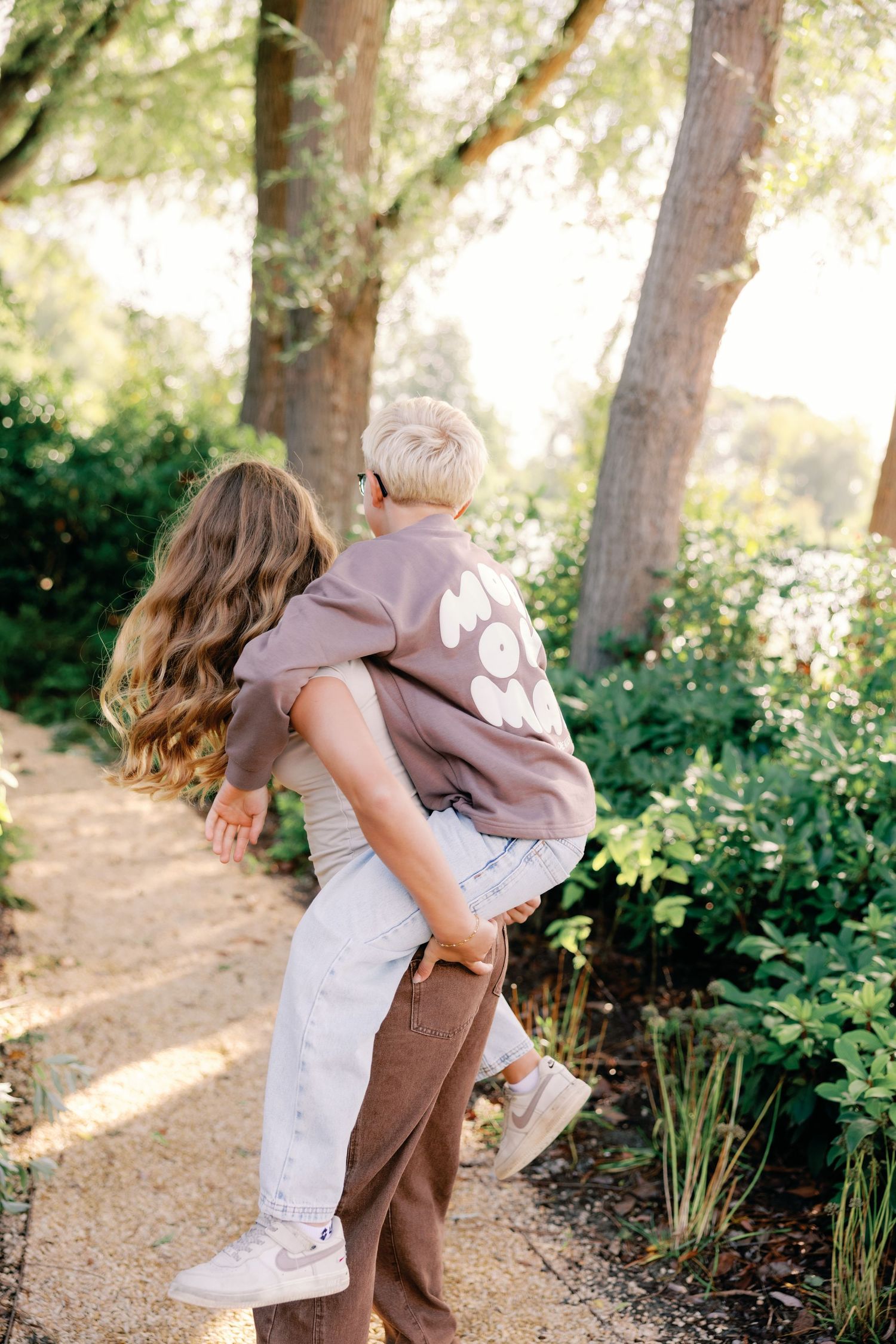 A girl with long wavy hair gives a piggyback ride to a blond boy in a brown sweatshirt on a sunlit garden path lined with trees.