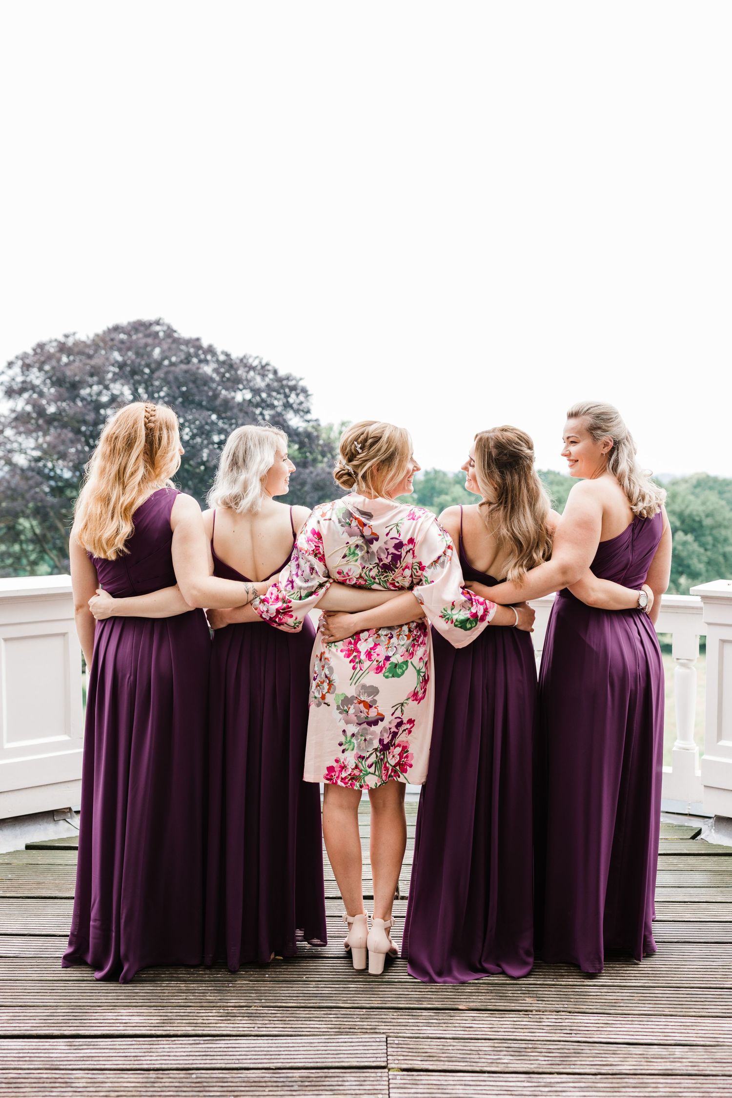 A group of bridesmaids in purple dresses and one in floral stand together with arms around each other on a deck.
