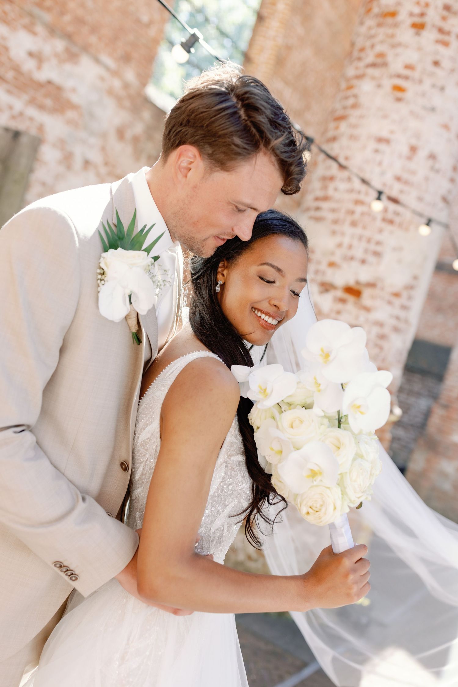 A tender wedding moment against a rustic brick wall with white floral bouquet.