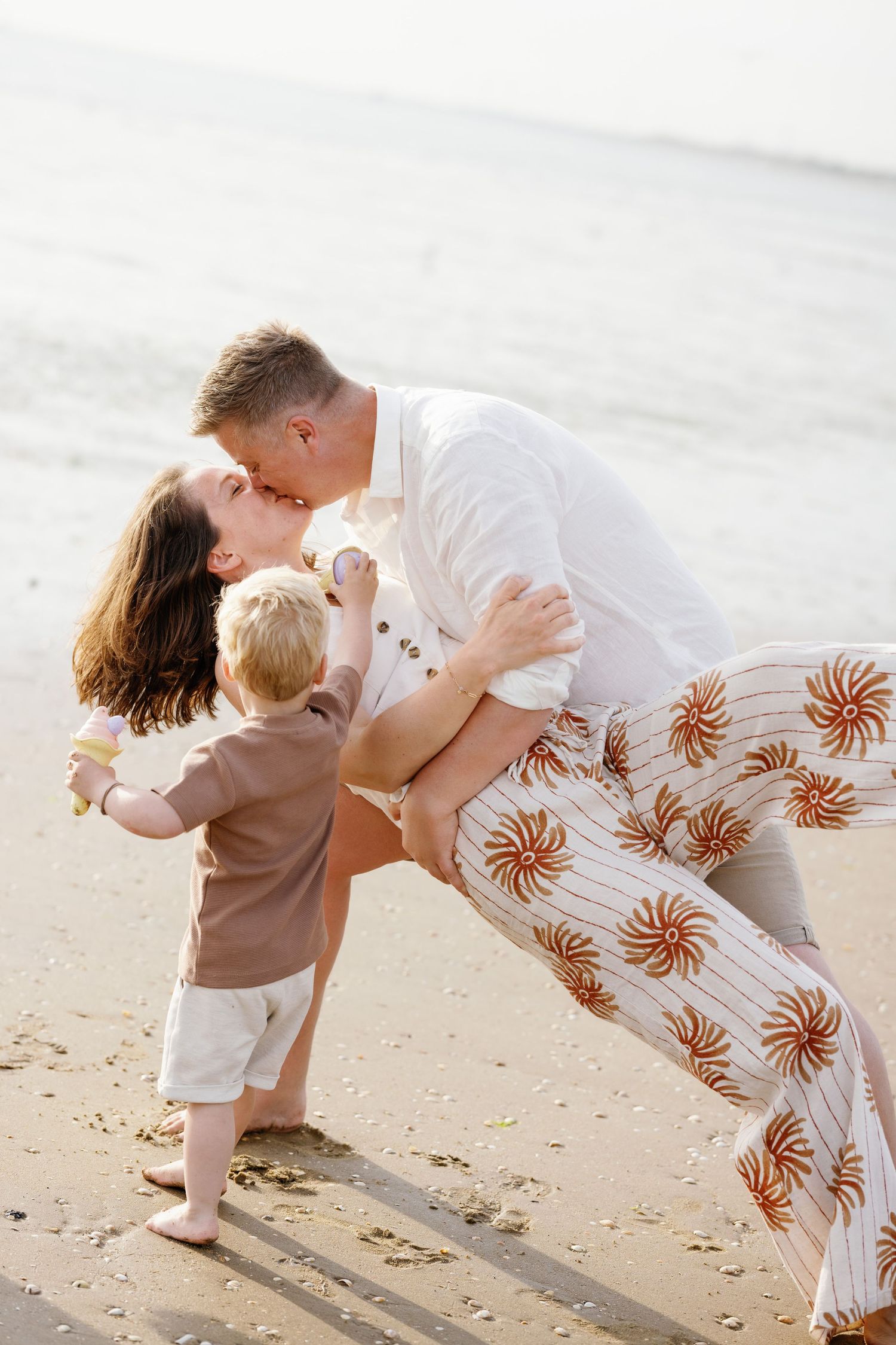 A couple shares a romantic dip kiss on the beach while their young toddler stands nearby holding a flower.
