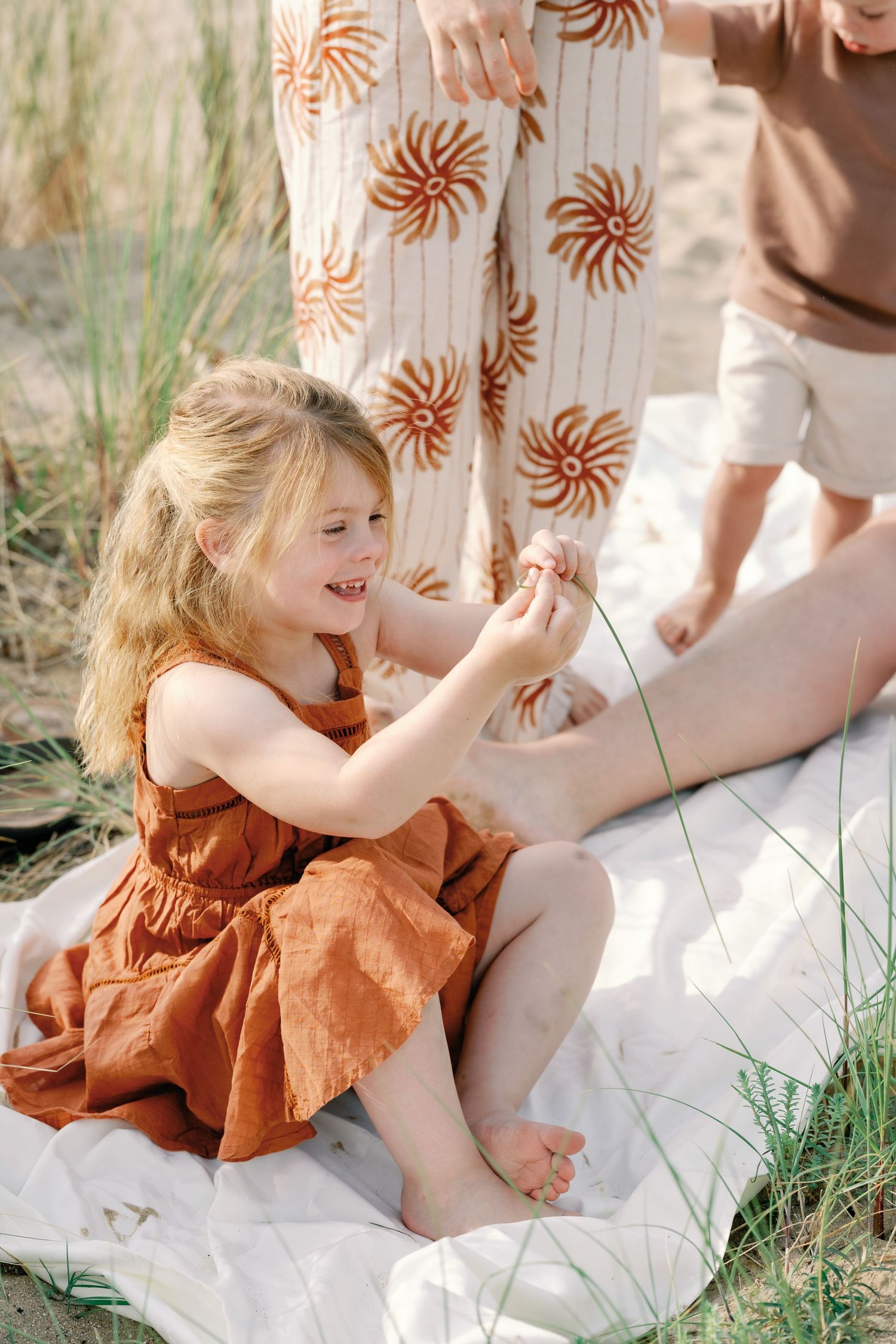 Young girl in rust dress sits barefoot on blanket outdoors, smiling and playing with a flower near family.
