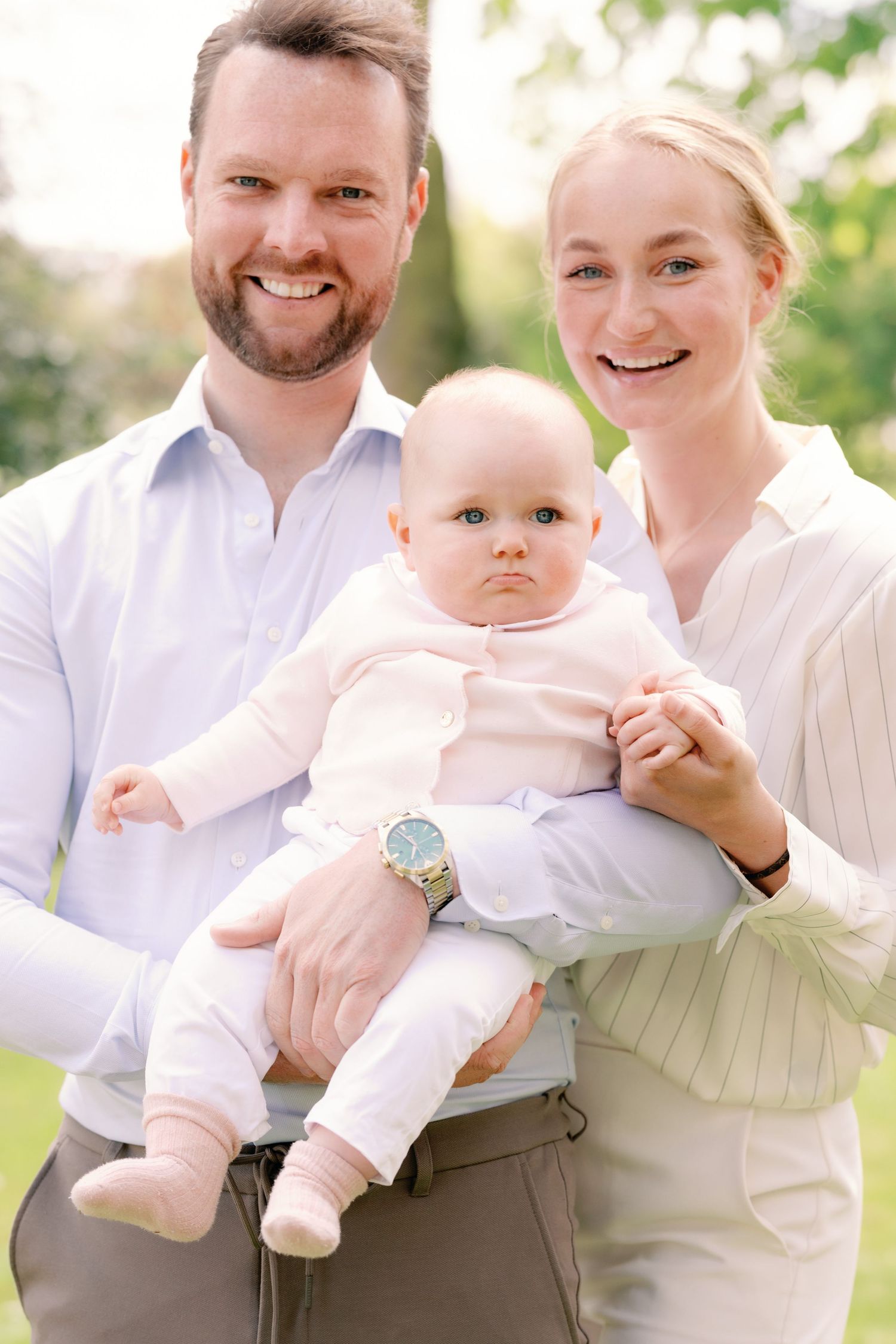Smiling couple holding their baby outdoors in a sunny green garden setting.