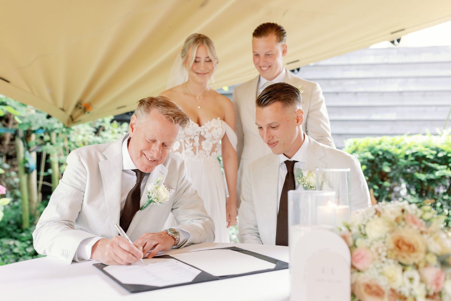 A wedding ceremony moment captured under a white tent as documents are being signed at an outdoor venue.