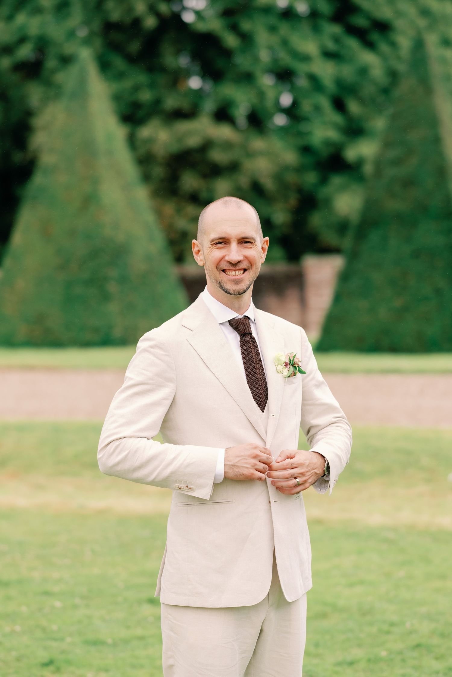 A person in a cream-colored suit stands outdoors on a green lawn during a wedding photoshoot.