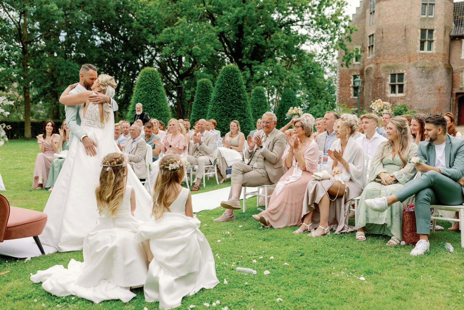 A group in light colored attire sits on chairs arranged in a semicircle on a grassy lawn near brick buildings.