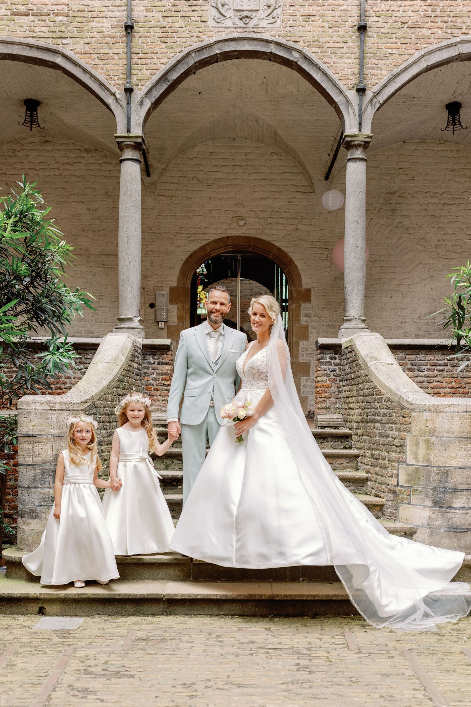 Wedding party poses under elegant stone archways in a historic courtyard setting with white gowns and light gray suits.