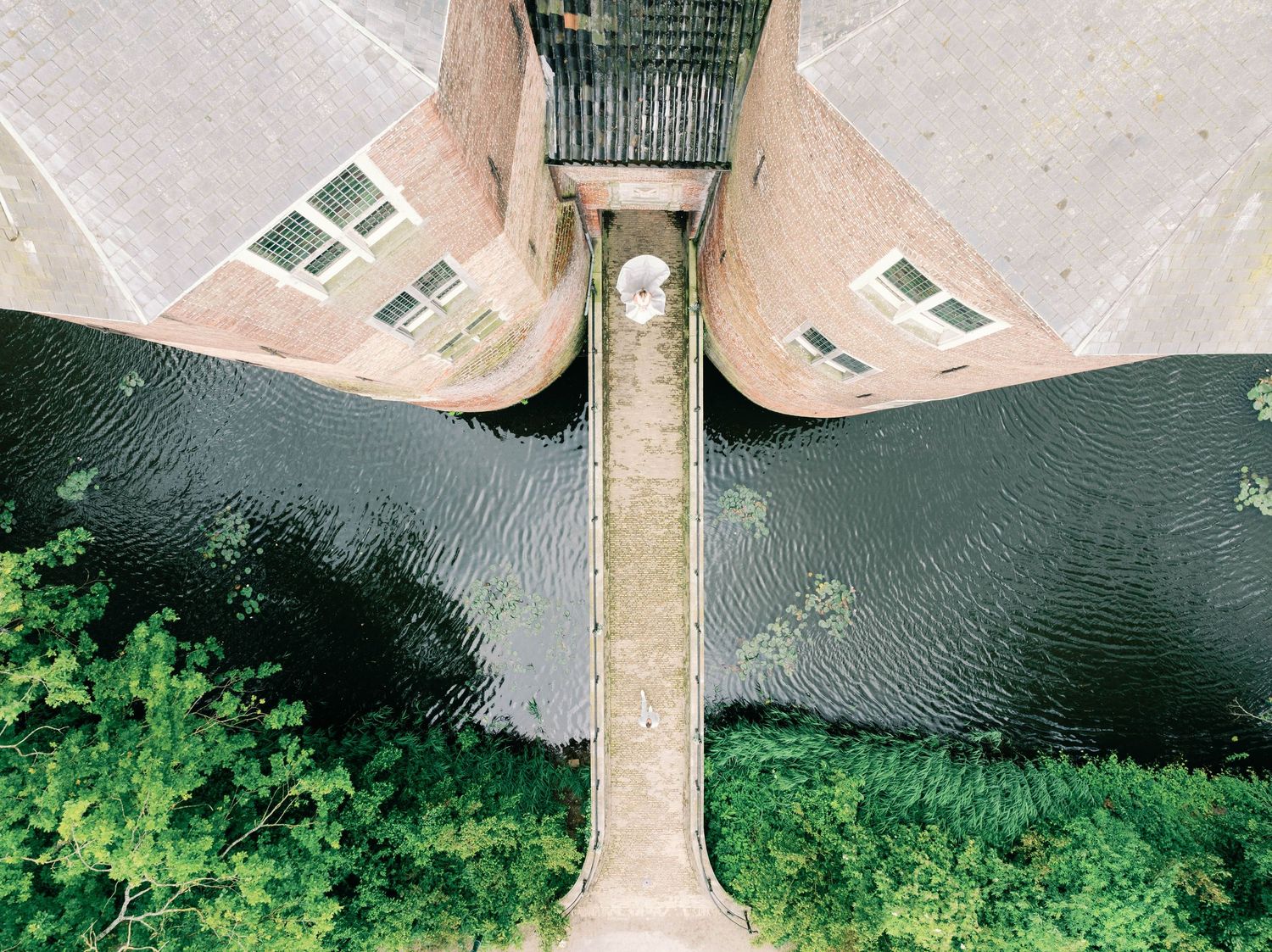 Aerial view of a tall tower casting shadow on dark water surrounded by green foliage and buildings.