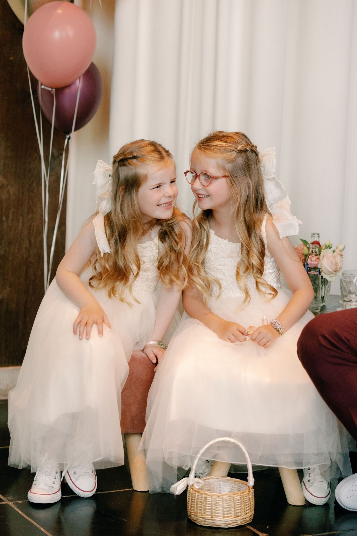 Two young girls in white dresses and pink balloons share joyful moments together during a special celebration.