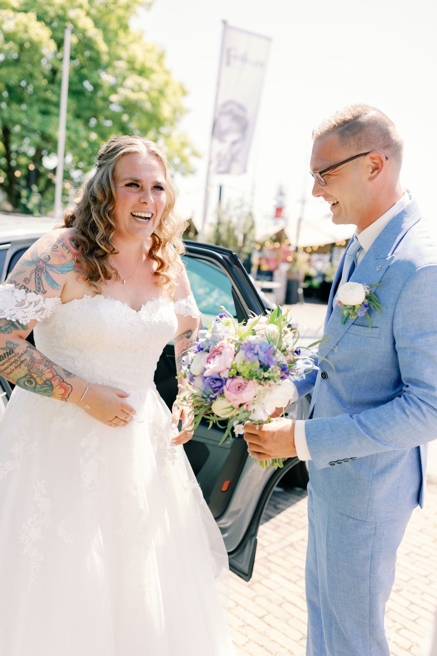 Bride in white lace gown and groom in light blue suit share a joyful moment with a colorful bouquet outdoors.
