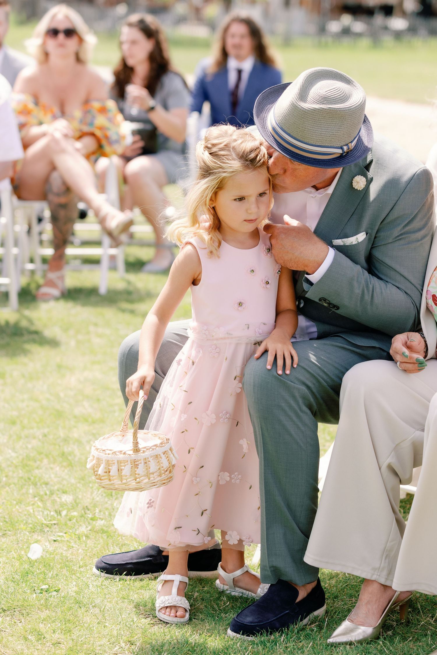 A young child in a pink dress sits with an adult in formal wear on grass during an outdoor gathering.