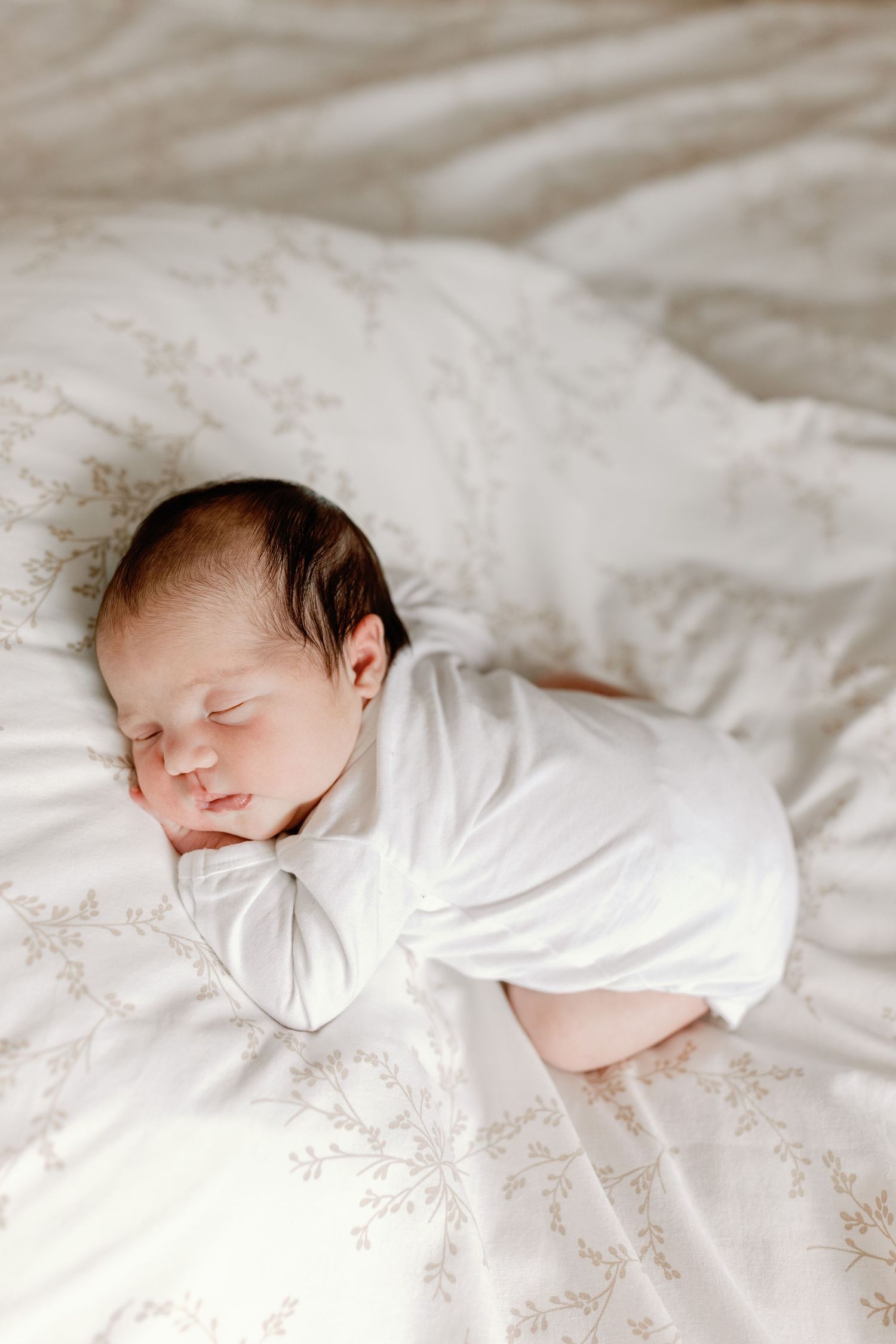 Sleeping newborn baby in white clothing rests peacefully on floral patterned bedding.